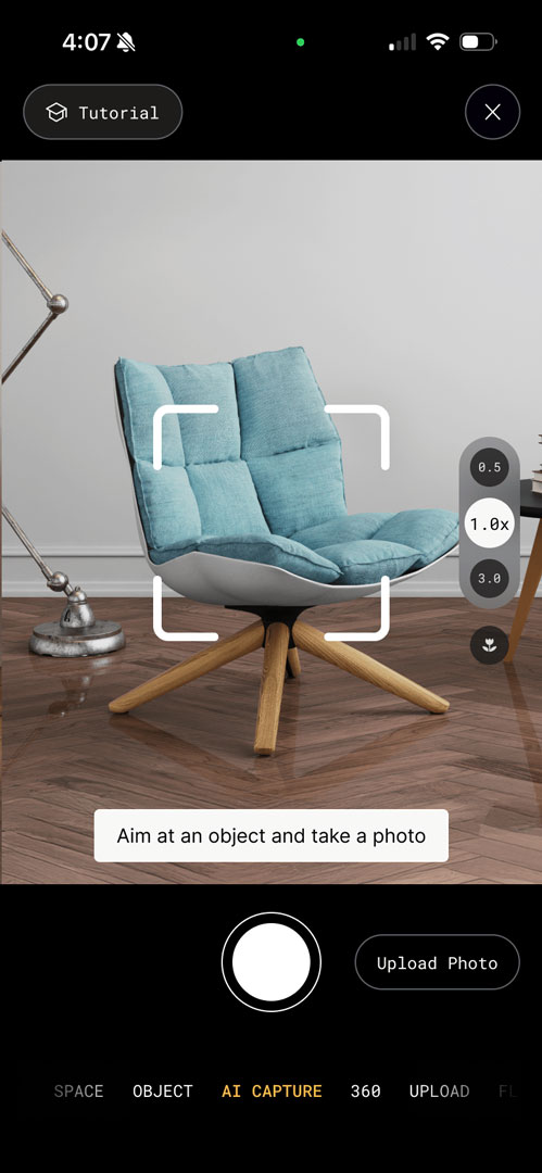 Polycam mobile app showing an in-session capture view of a light blue upholstered chair with wooden legs on a hardwood floor. The interface displays a white viewfinder frame around the chair with the instruction "Aim at an object and take a photo" at the bottom.