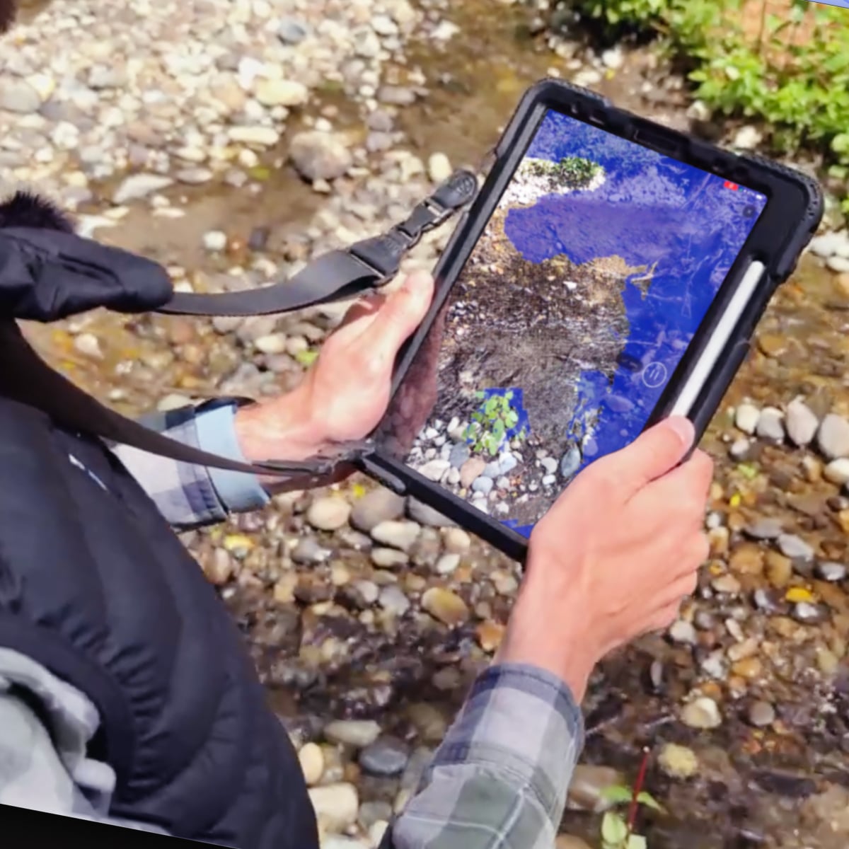 An engineer holds a rugged tablet running Polycam software while scanning a creek bed. The device displays a 3D scan visualization of the terrain.