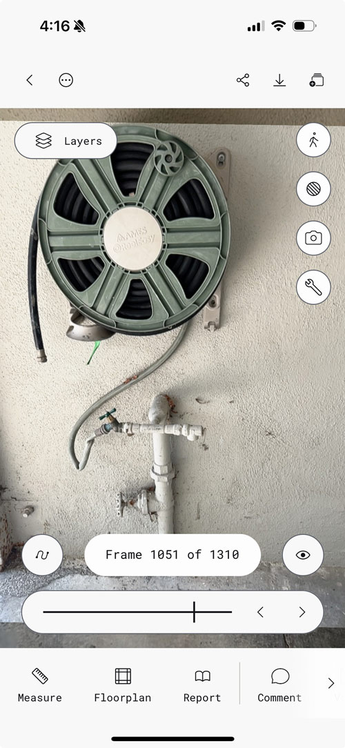 Polycam walkthrough mode interface showing frame 1051 of 1310, featuring a close-up view of a sage green garden hose reel mounted on a textured white wall above white PVC plumbing pipes. The mobile interface displays navigation controls, a Layers button, and bottom menu with Measure, Floorplan, Report, and Comment options.