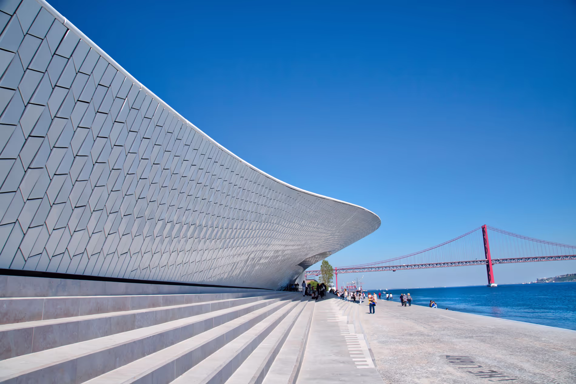 People walking along wide steps and modern curved building facade beside a river with a large red suspension bridge in the background under a clear blue sky.