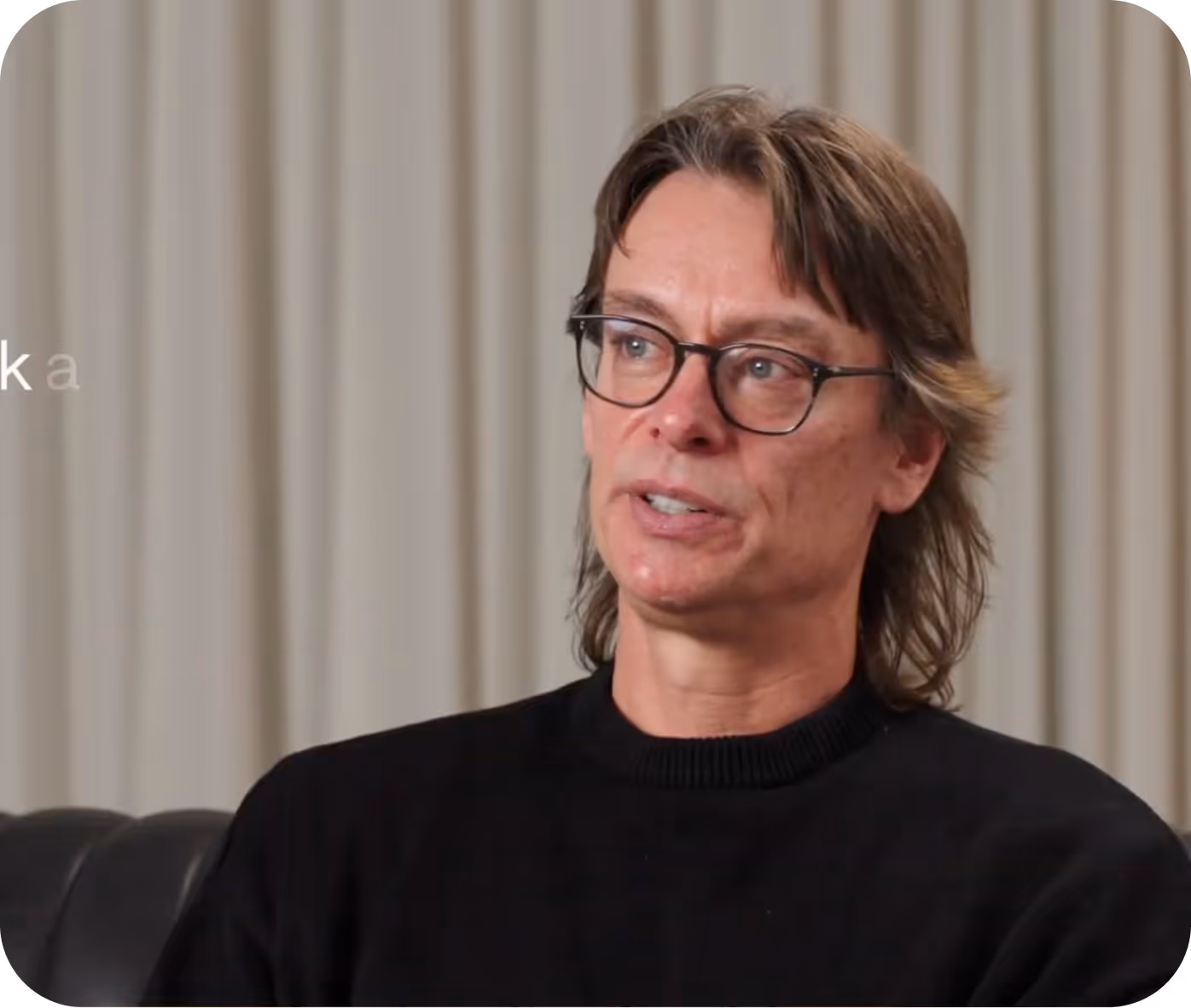 Middle-aged man with glasses and medium-length brown hair wearing a black sweater, sitting on a dark leather couch with beige curtains in the background.