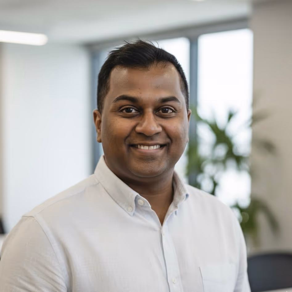 Smiling man wearing a white button-up shirt in a bright office with blurred windows and plants in the background.