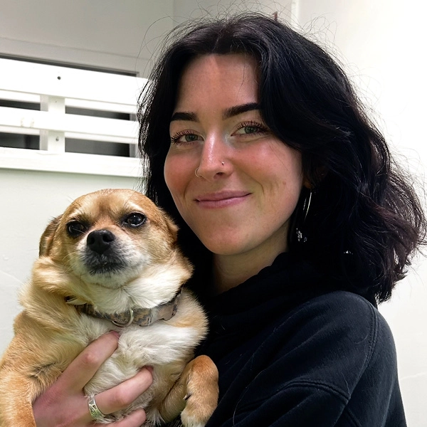 A photo of Brooklyn smiling at the camera with her small brown short-haired dog indoors.