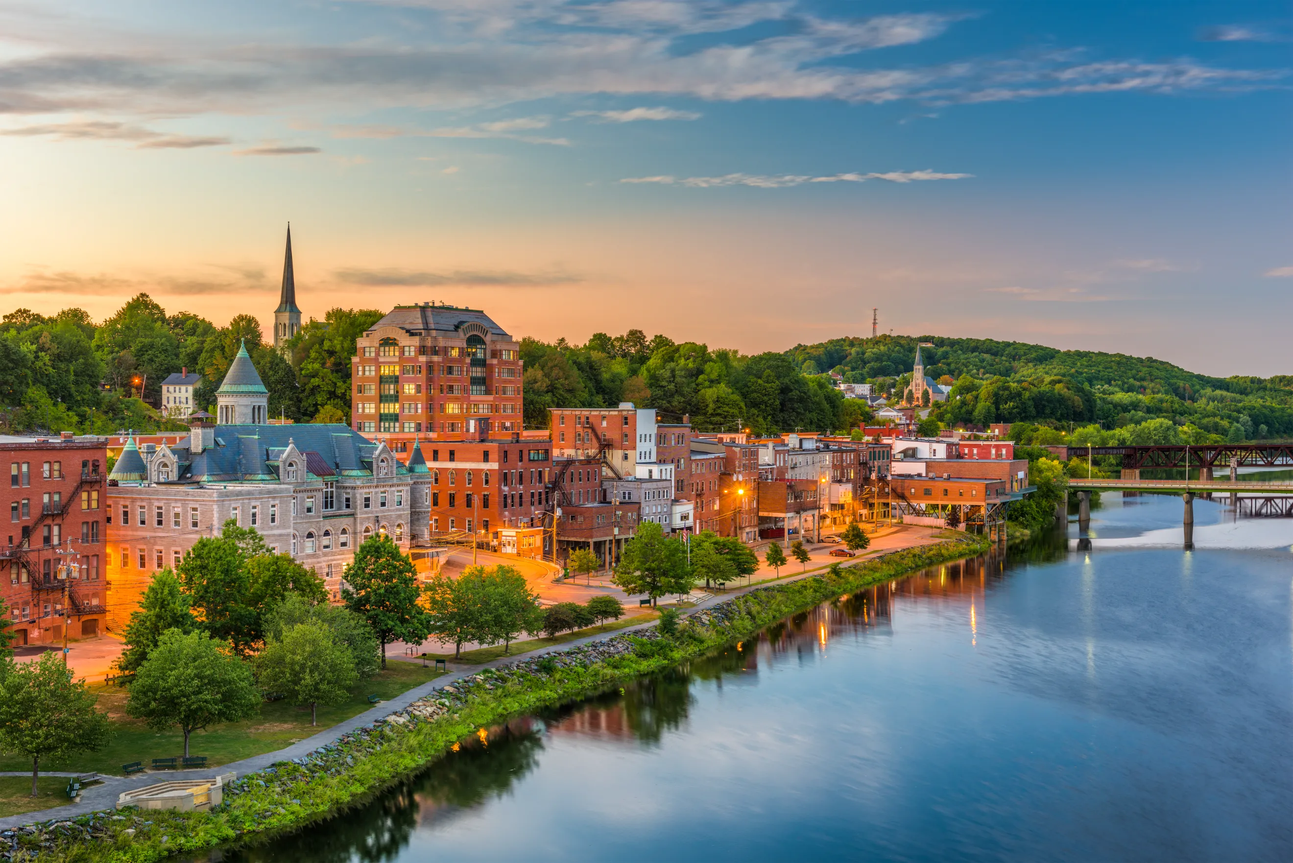 A charming riverside town in Maine at dusk, featuring historic brick buildings, a church steeple, and a calm waterfront reflecting the warm glow of streetlights.