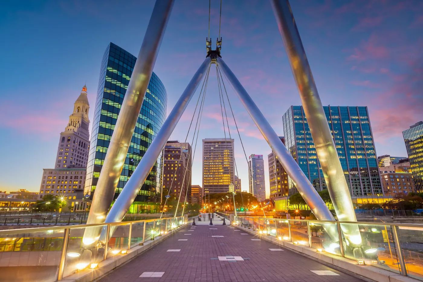 A vibrant cityscape in Hartford, Connecticut, showcasing a modern pedestrian bridge leading to illuminated office buildings under a colorful evening sky.