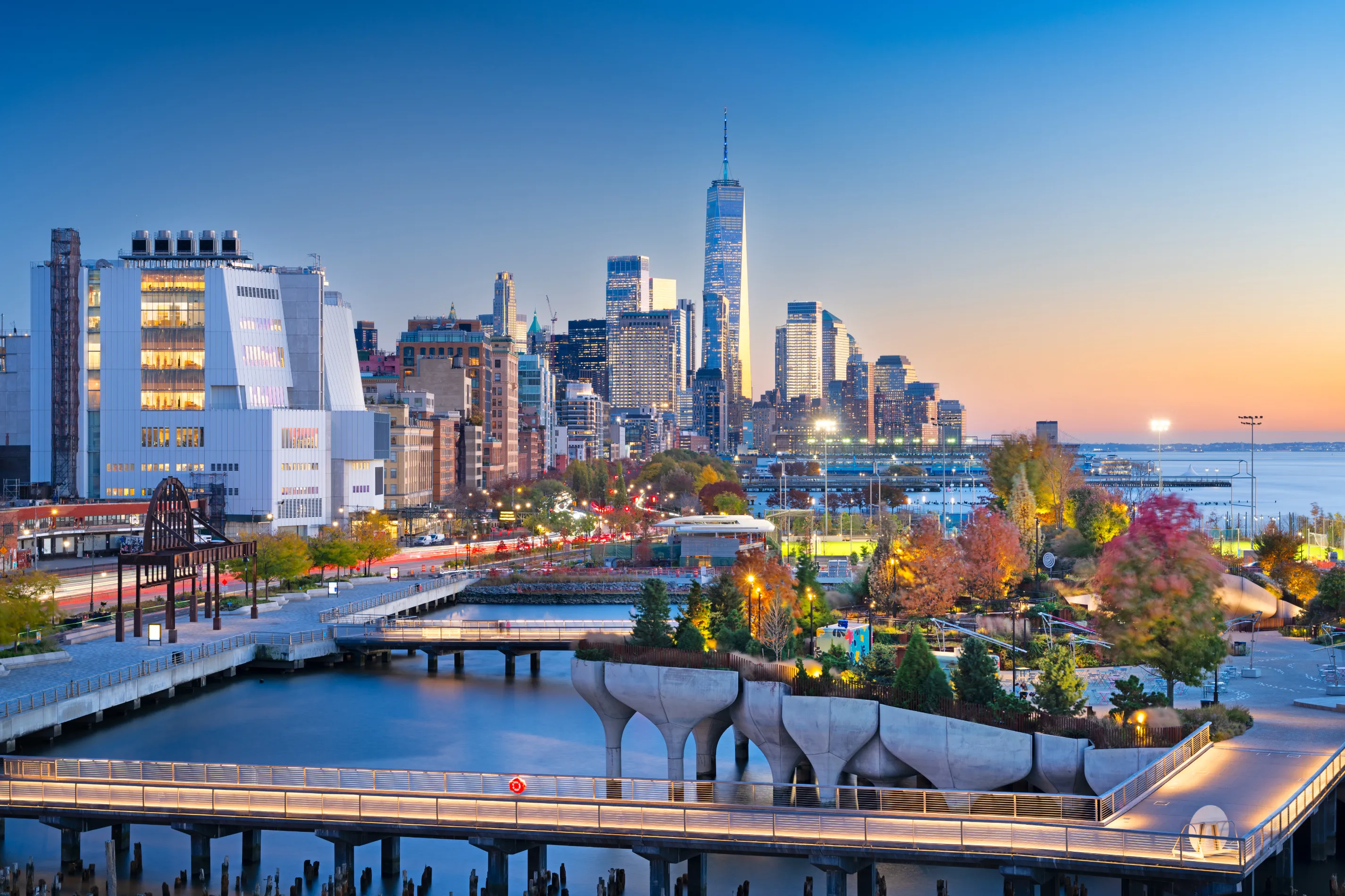 A stunning view of the New York City skyline at sunset, featuring the Hudson River, modern skyscrapers, and the iconic One World Trade Center.