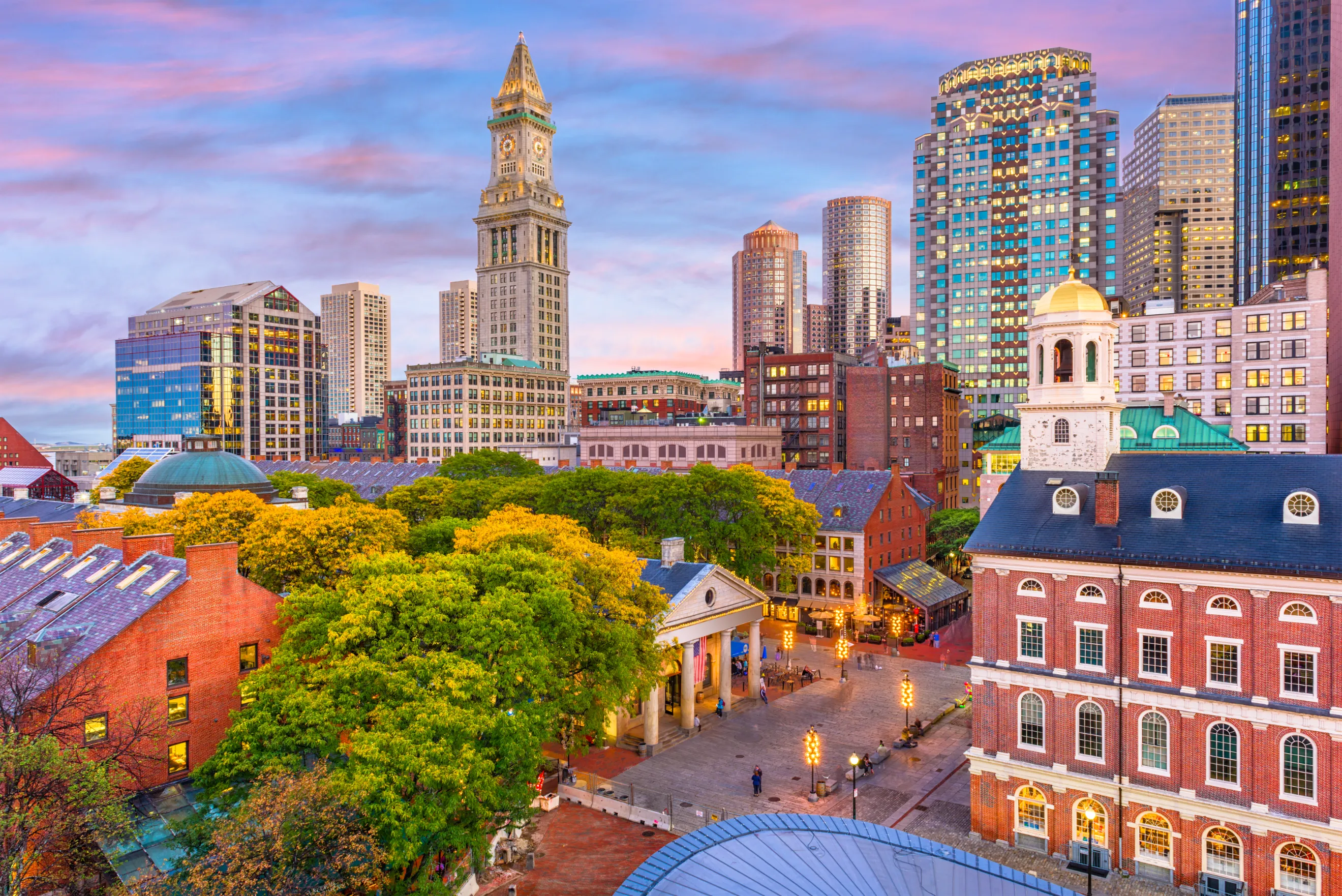 A lively Boston cityscape at dusk, featuring Faneuil Hall, historic buildings, and modern skyscrapers illuminated under a pastel-colored sunset sky.