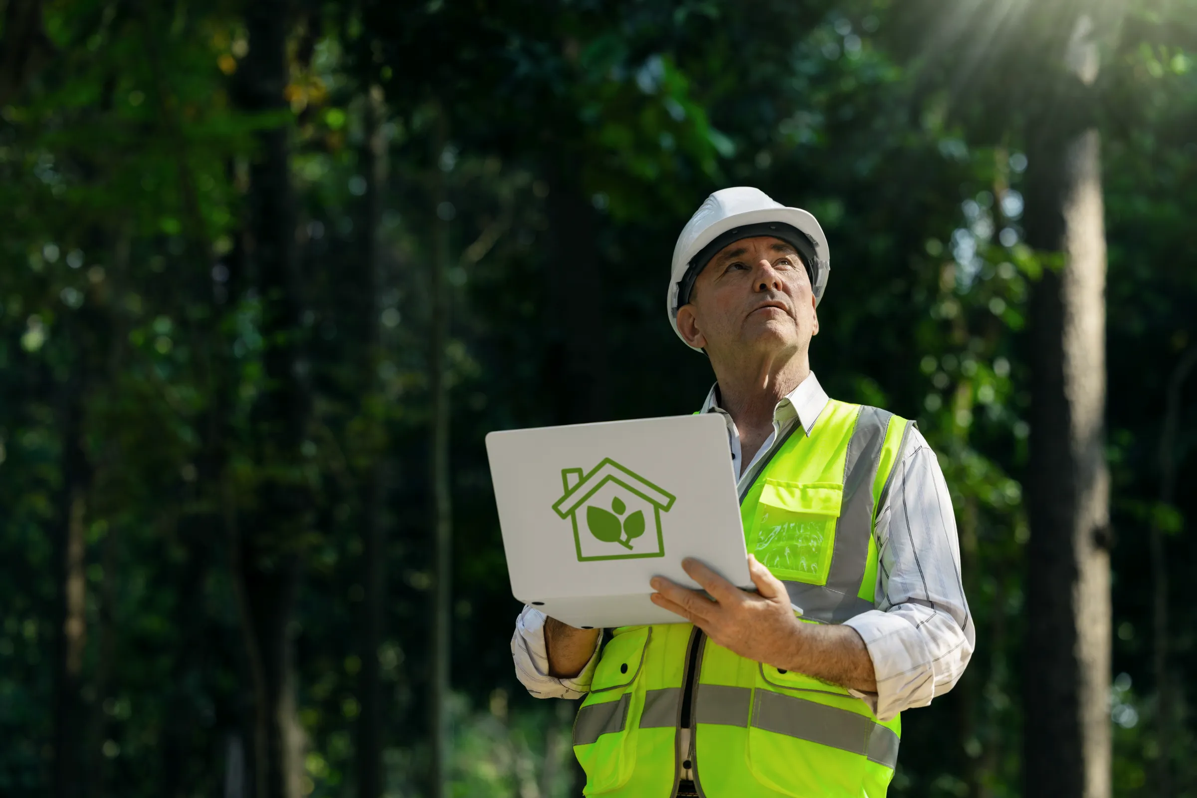Construction worker wearing a hard hat and safety vest, holding a laptop with a green building logo, standing amidst a lush forest background.
