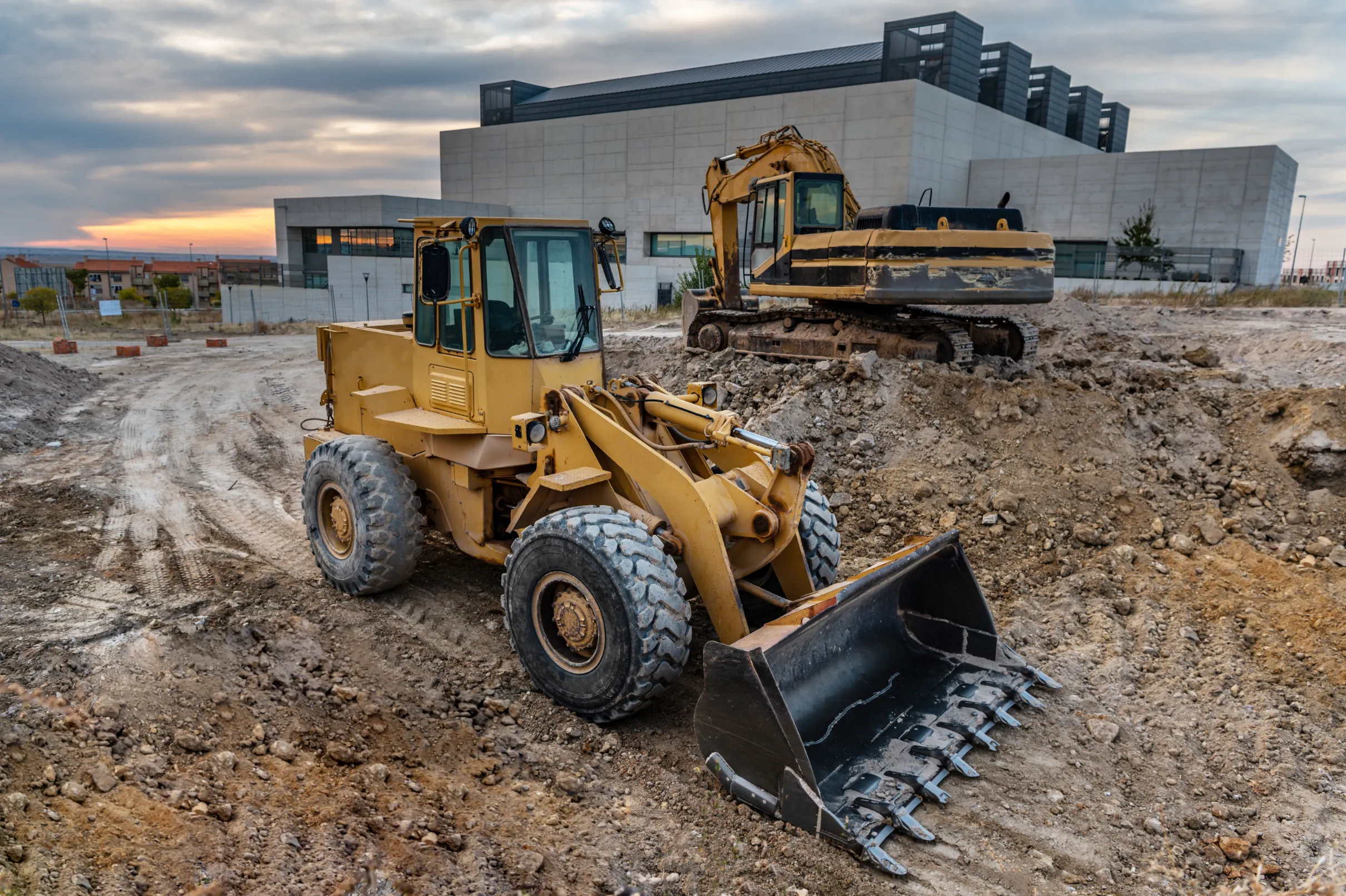 Two heavy construction vehicles, including a loader and excavator, stationed on a dirt construction site with a modern building in the background during sunset.