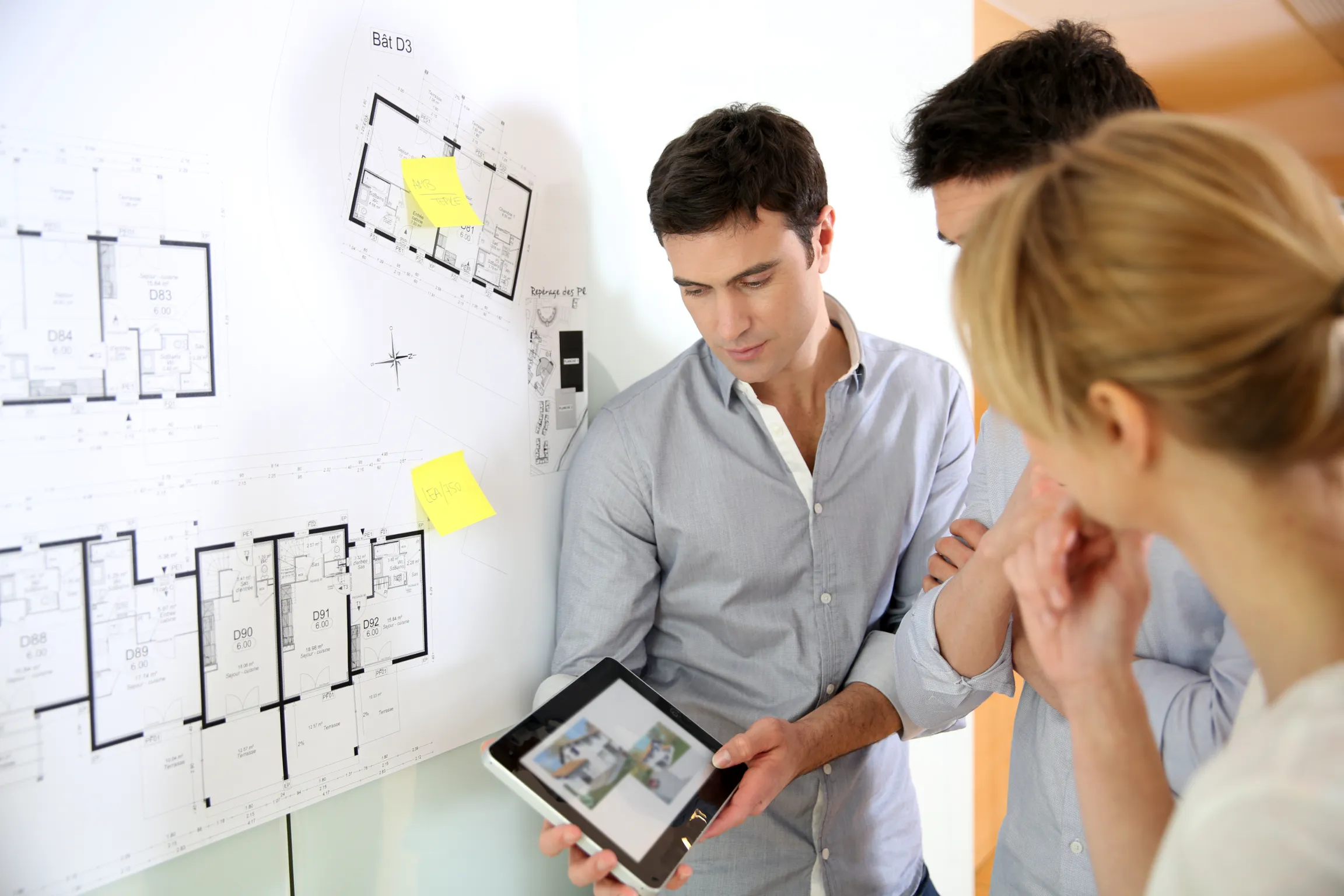 Architects and designers discussing building plans pinned to a wall, using a tablet to review construction project details in a well-lit office space.