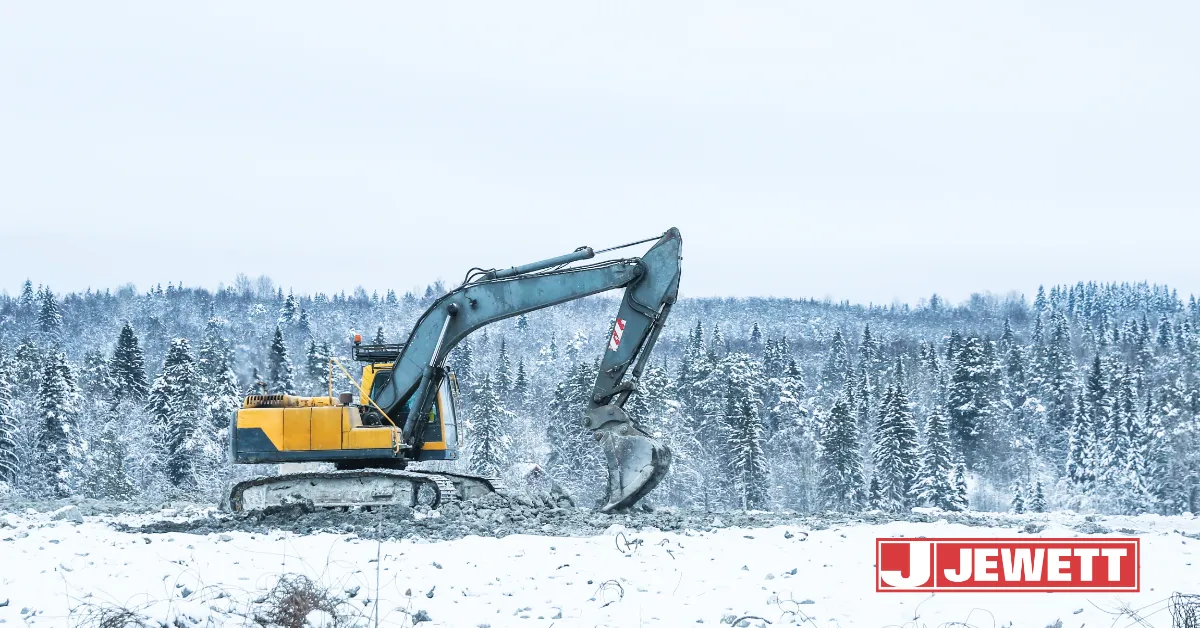 Excavator operating on a snowy commercial construction site during winter, with snow-covered trees in the background.