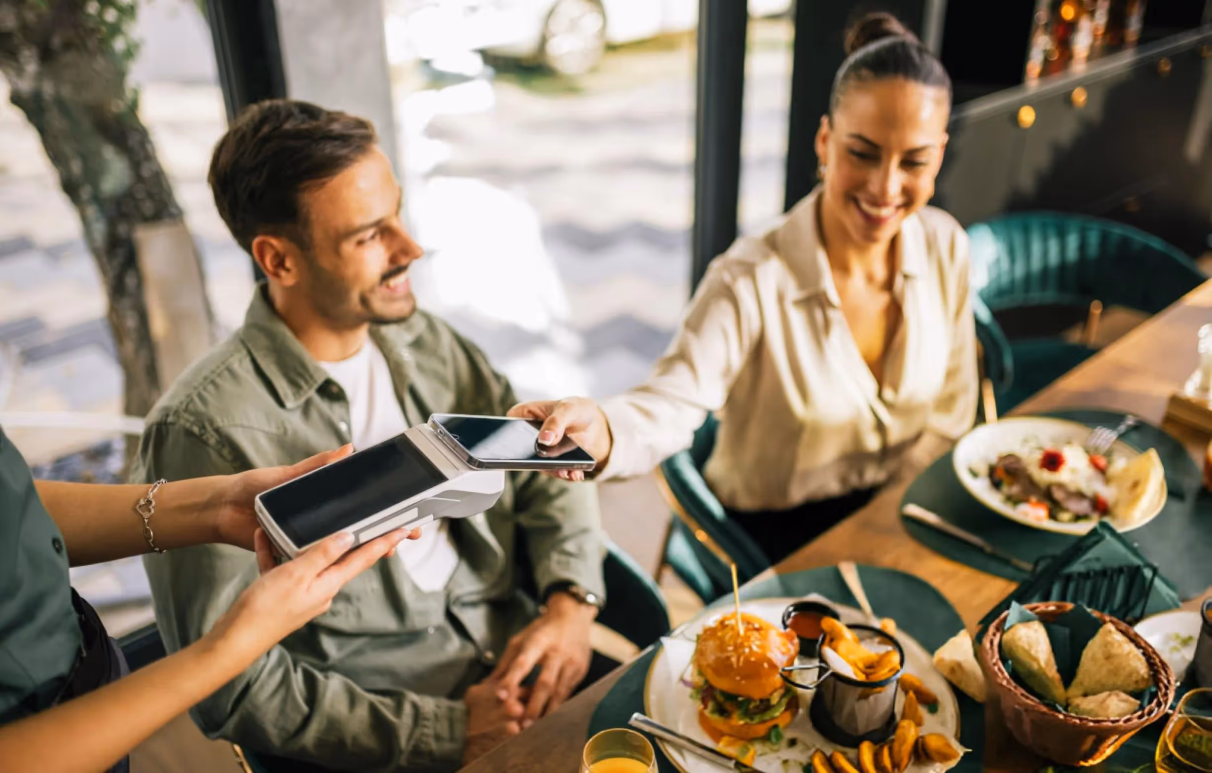 Une femme souriante paie avec son téléphone à un serveur dans un restaurant, un homme souriant est assis à côté d'elle avec des plats devant eux.