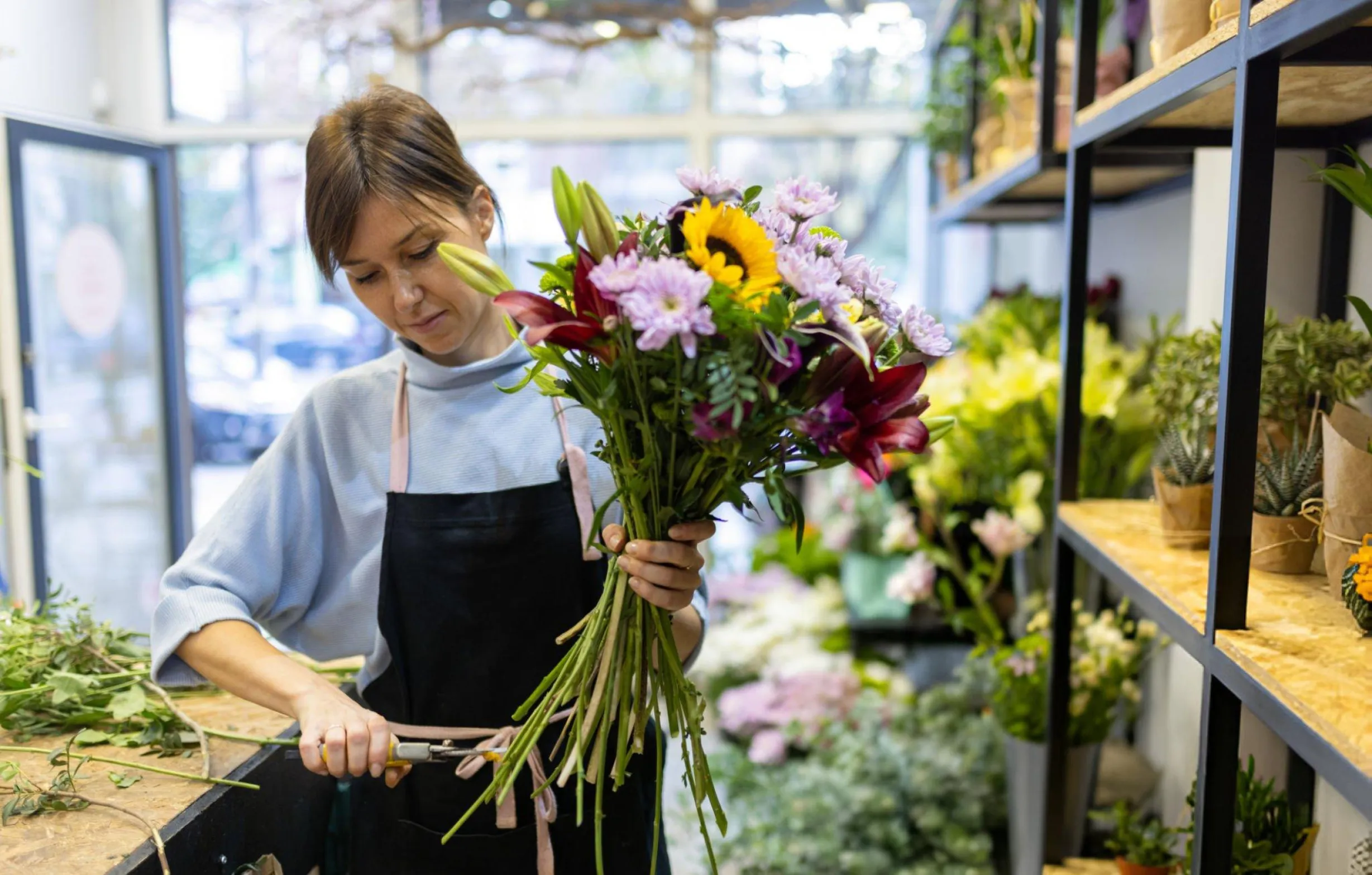 Femme en tablier noir préparant un bouquet de fleurs colorées dans une boutique de fleurs lumineuse.