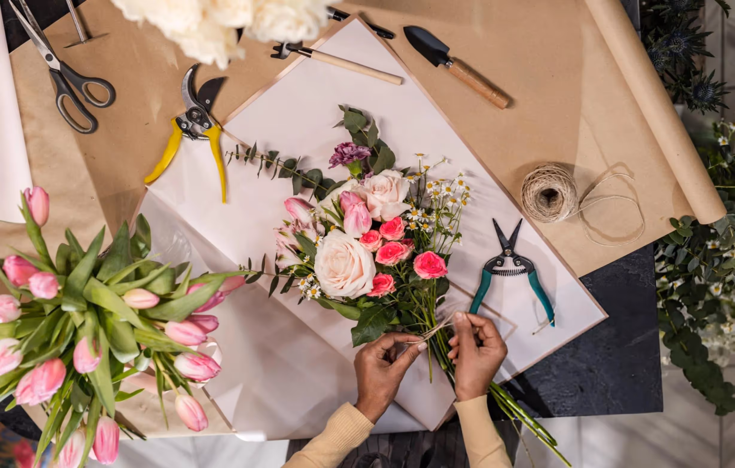 Une personne prépare un bouquet de fleurs avec des roses roses, des tulipes et des fleurs sauvages, entouré d'outils de jardinage et de rouleaux de papier kraft.