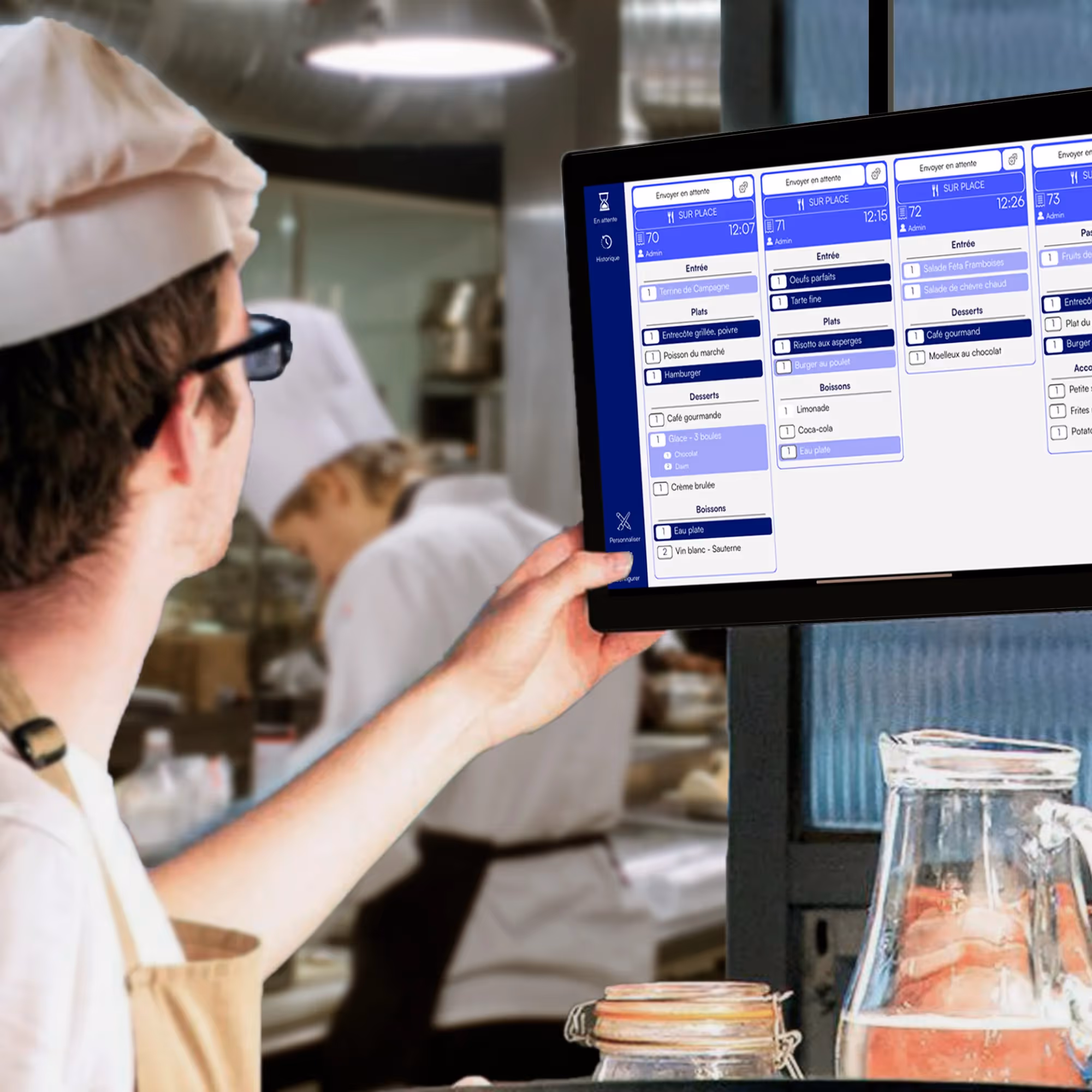 Un chef cuisinier regardant un écran tactile affichant des commandes de plats et boissons dans une cuisine professionnelle.