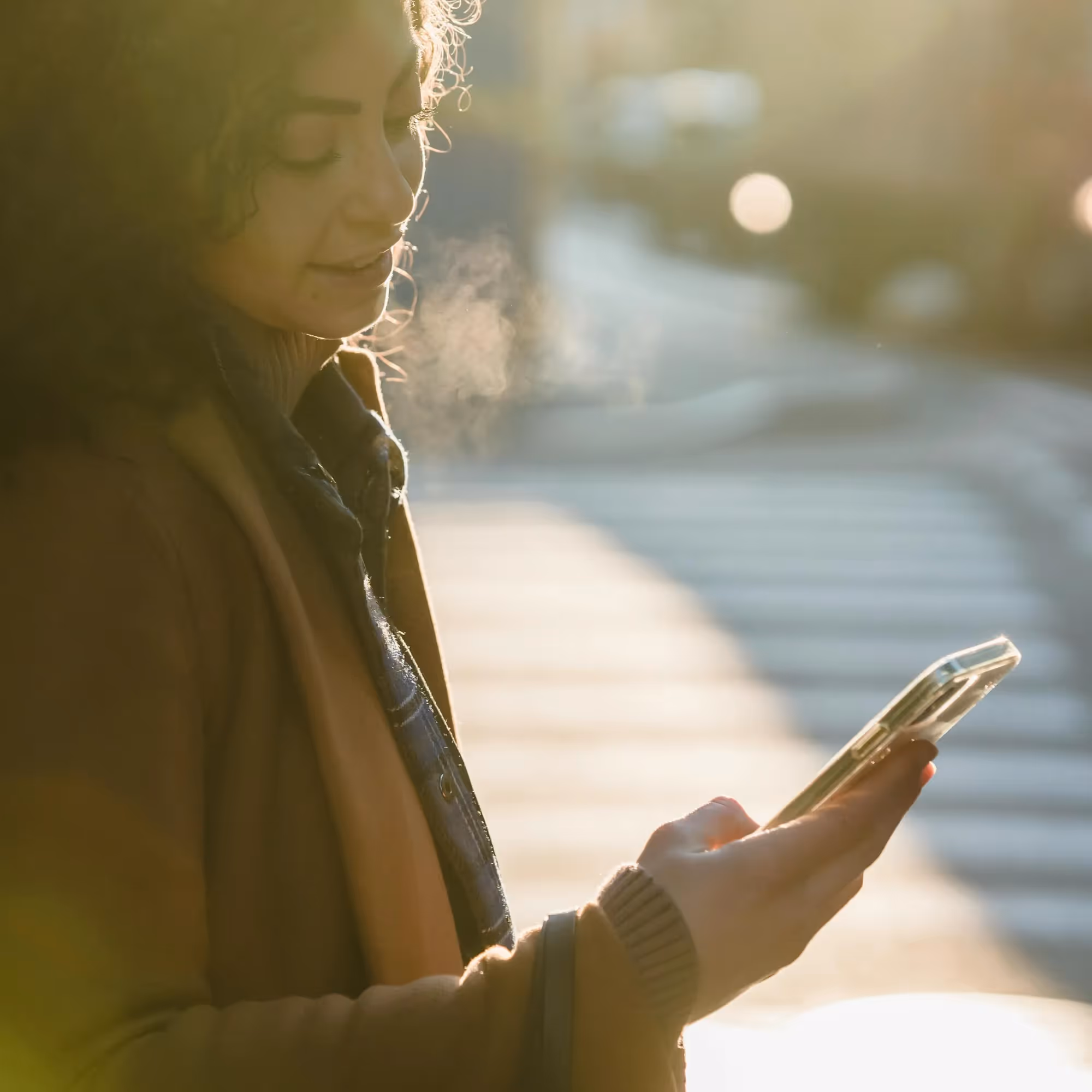 Une femme souriante portant un manteau regarde son téléphone en plein air par une journée ensoleillée.