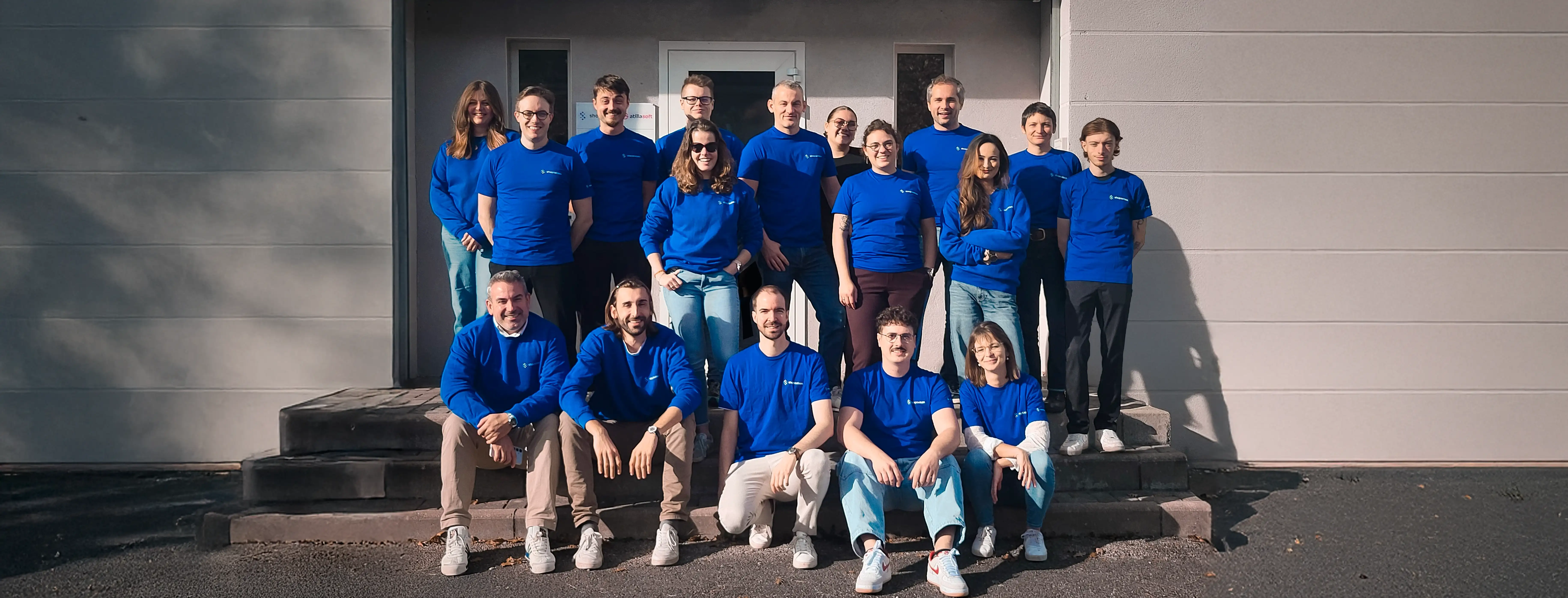 Groupe de 19 personnes portant des tee-shirts bleus posant devant un bâtiment avec marche.