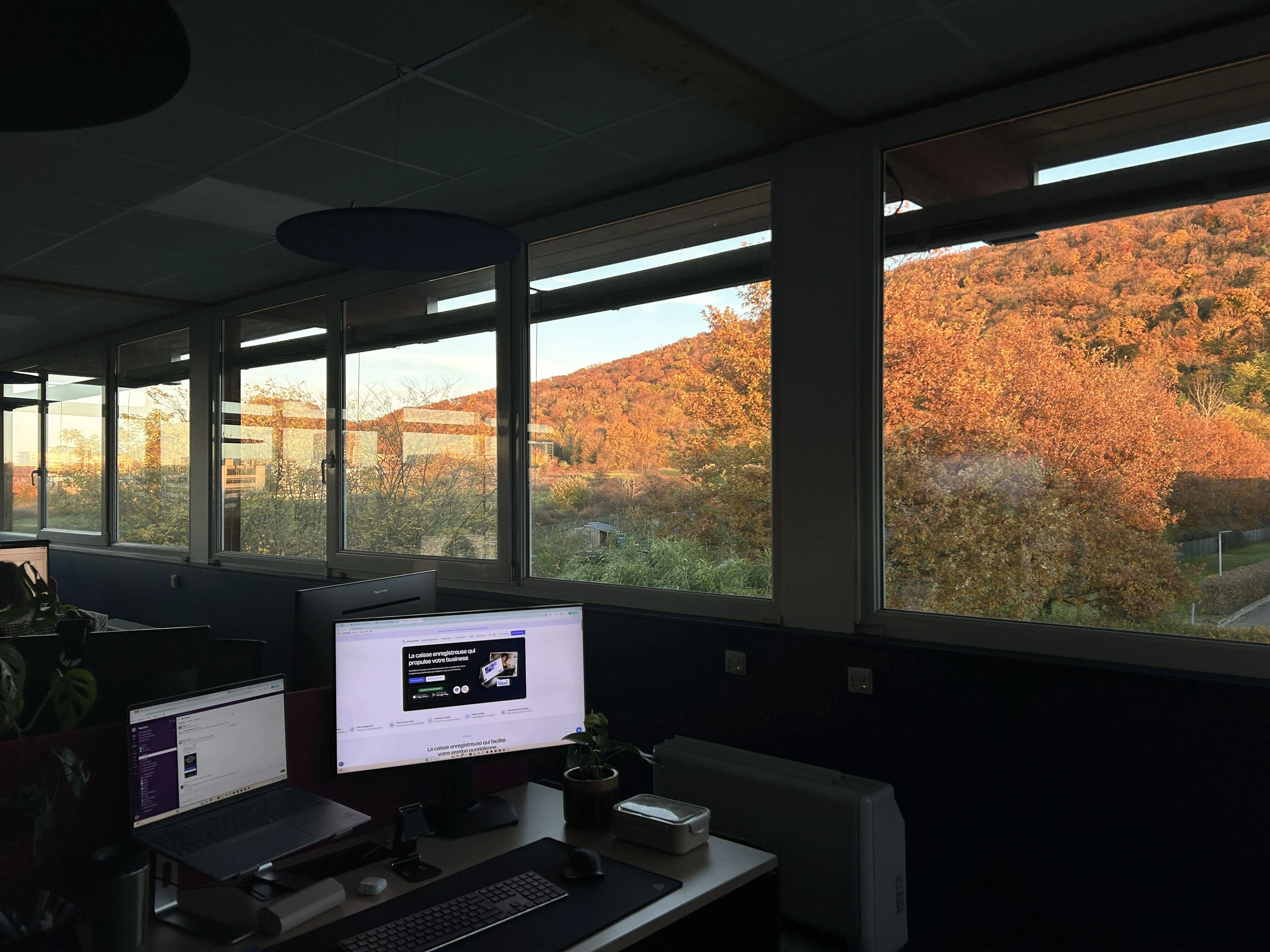 Bureau avec ordinateur portable et écran, grande fenêtre montrant arbres aux feuilles orange d'automne et colline sous un ciel bleu clair.