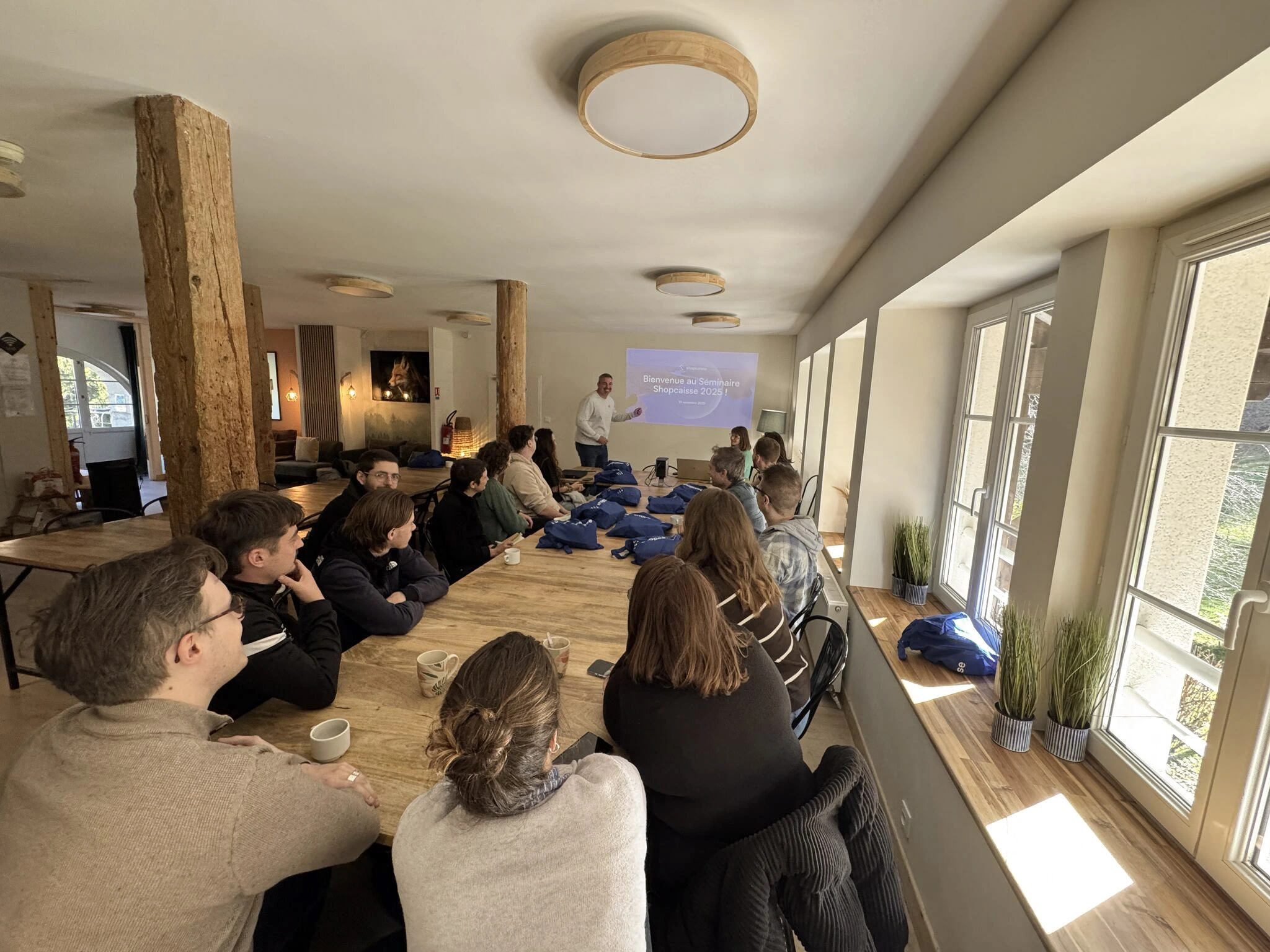 Groupe de personnes assises autour d'une grande table en bois, écoutant une présentation projetée murale avec le texte « Bienvenue au Séminaire Shopcaaisse 2025 ! » dans une salle lumineuse avec fenêtres et décorations végétales.