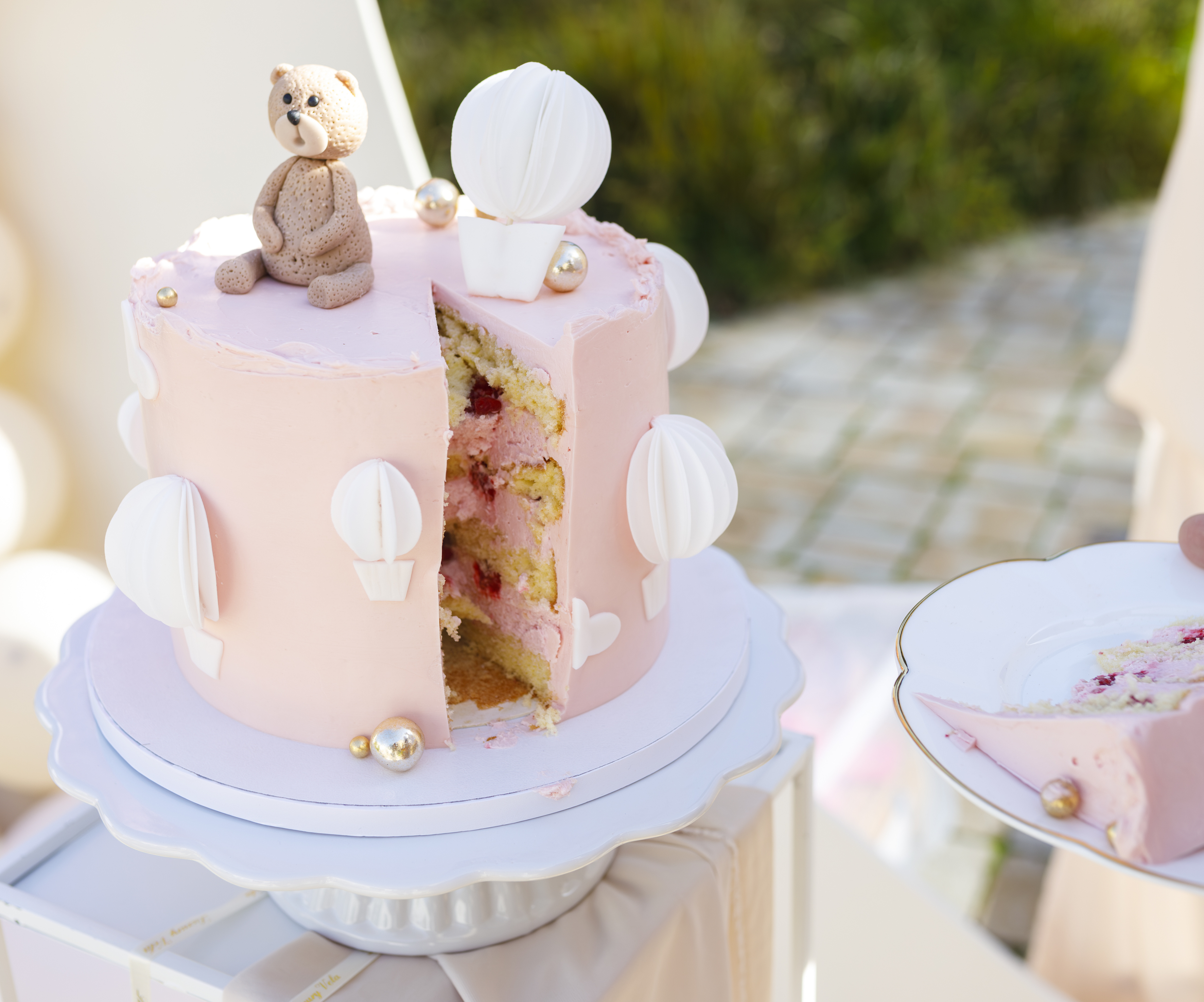 Rosa Babyshower-Torte mit Bärenfigur und Heißluftballon-Dekoration, mit Beeren-Füllung, serviert auf weißer Etagere im Freien