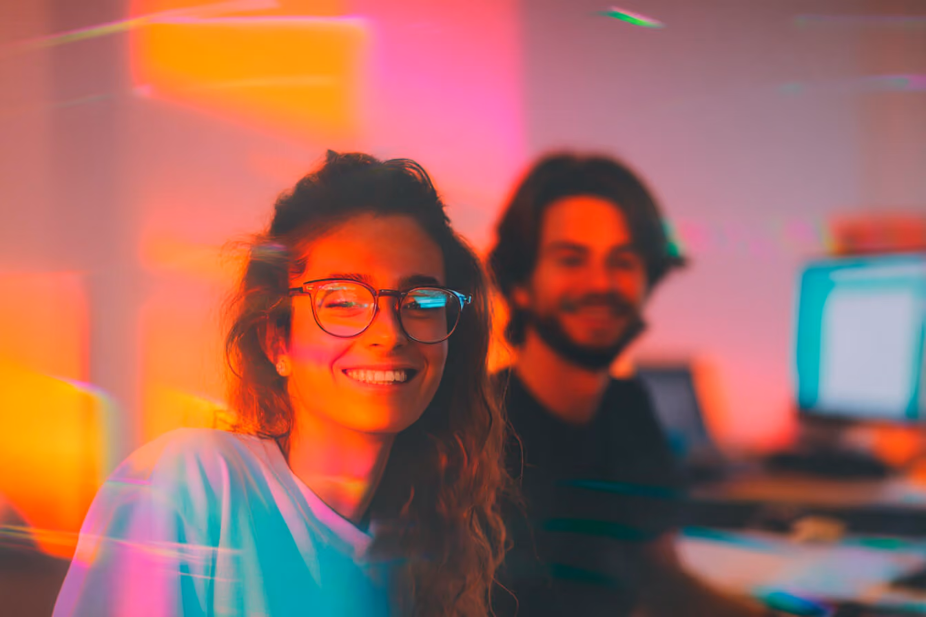 Smiling woman with glasses in colorful light, man with beard blurred in background in office setting.