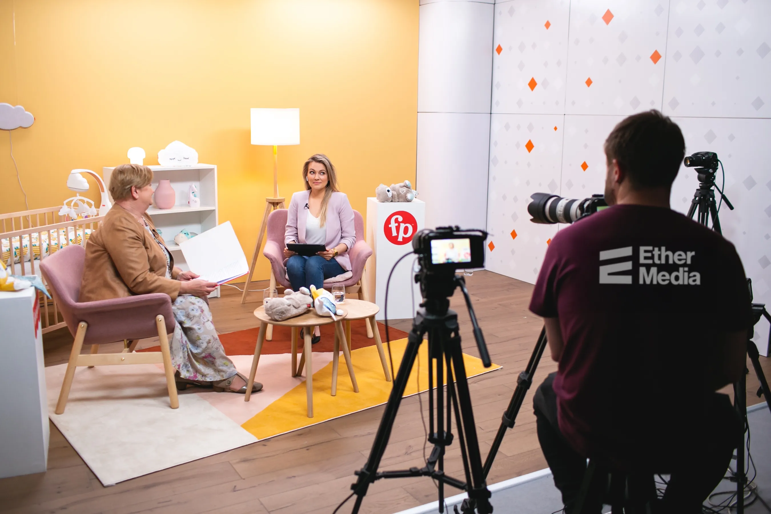 Dwóch kobiet rozmawia na tle żółtej ściany w studio nagraniowym z kamerzystą rejestrującym ich rozmowę.