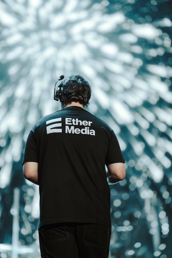 Mężczyzna z tyłu w czarnej koszulce z logo Ether Media i zestawem słuchawkowym na głowie na tle rozmytych świateł scenicznych.