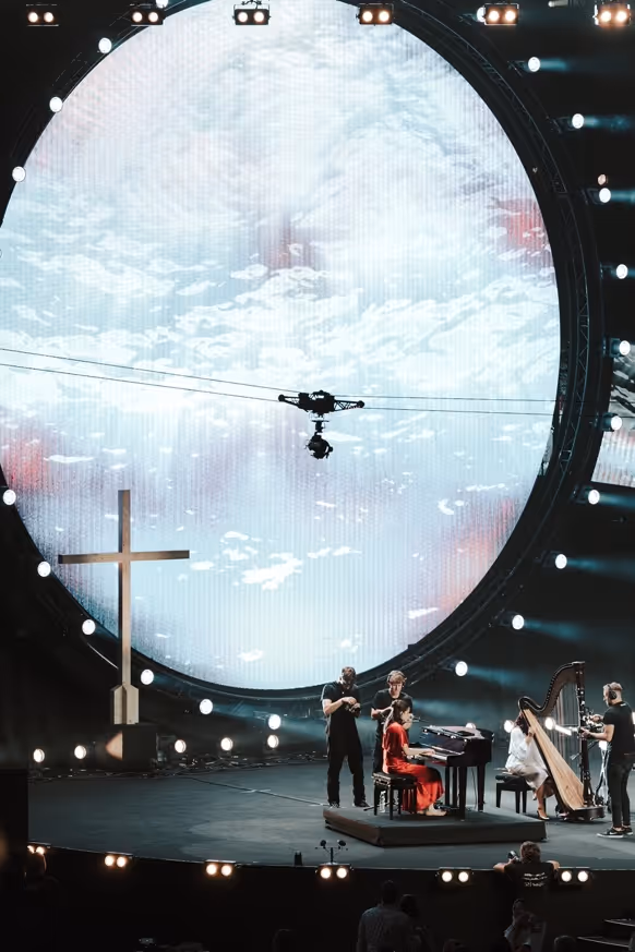 A woman in a red dress plays piano on stage with a harpist, next to a large illuminated cross and a big circular screen displaying clouds.