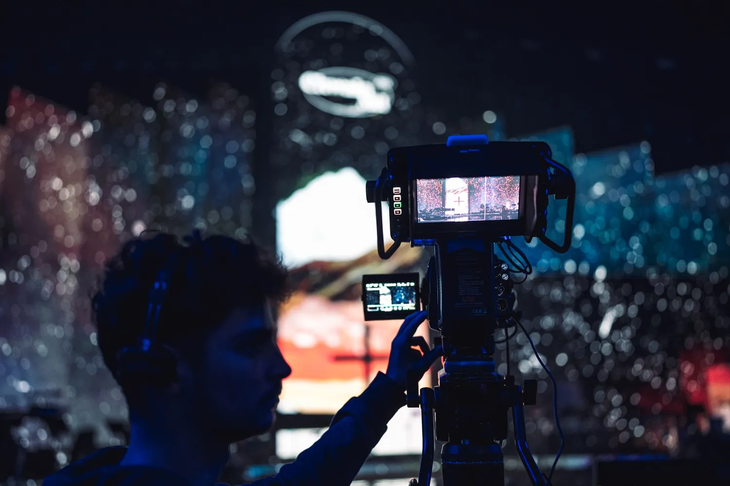 Operator adjusts professional video camera filming a vibrant stage with colorful lights in a dark setting.