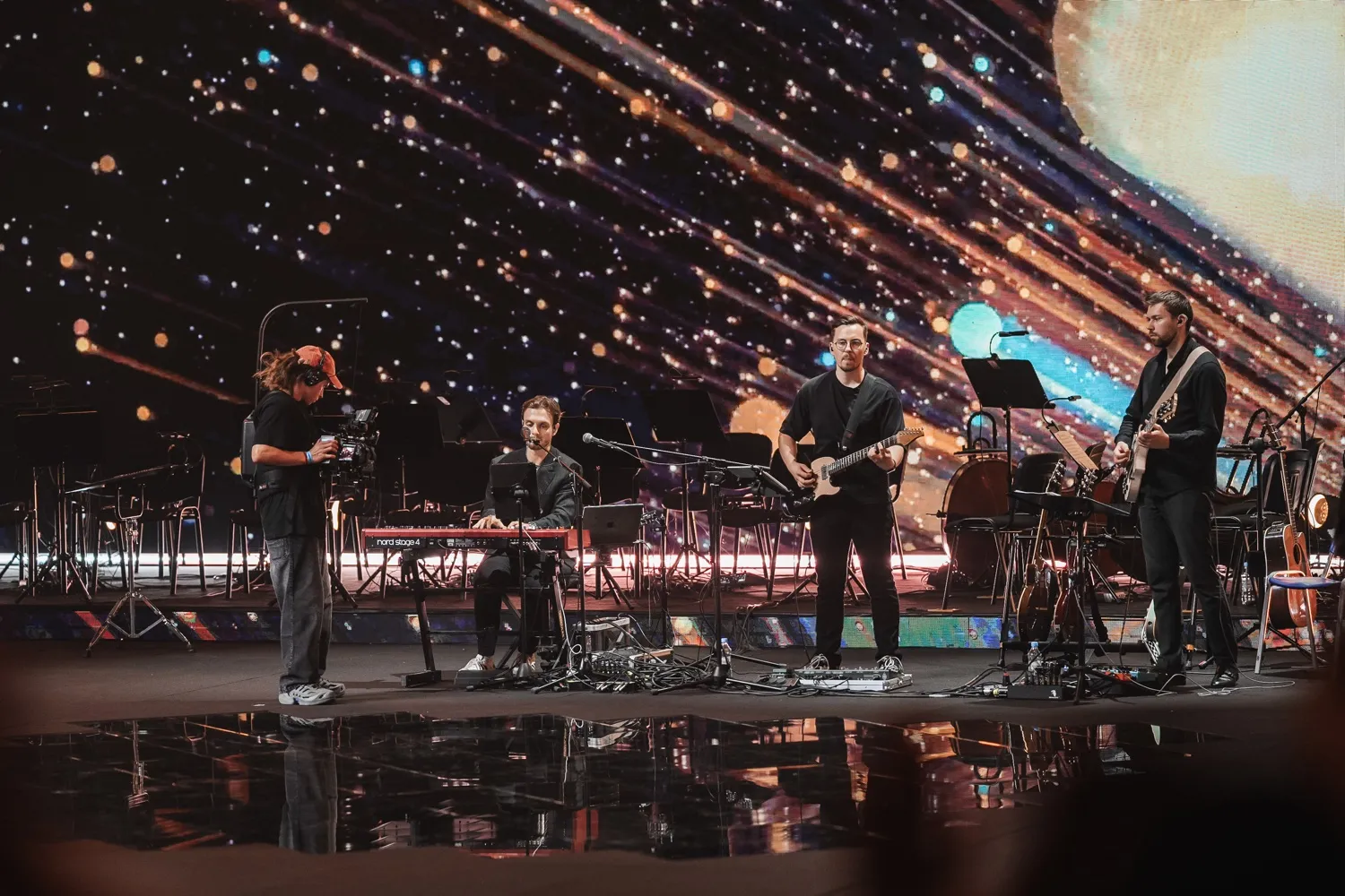 Three musicians on stage with guitars and keyboard, a cameraman filming, against a bright cosmic background.