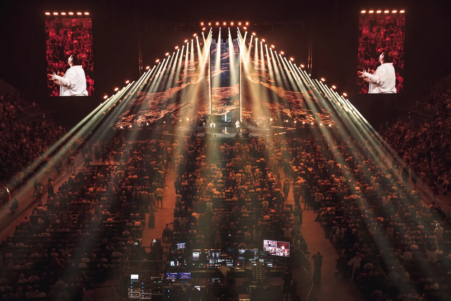 Widok na salę koncertową z dużą publicznością, sceną oświetloną mocnym światłem i dwoma ekranami bocznymi pokazującymi wykonawcę.
