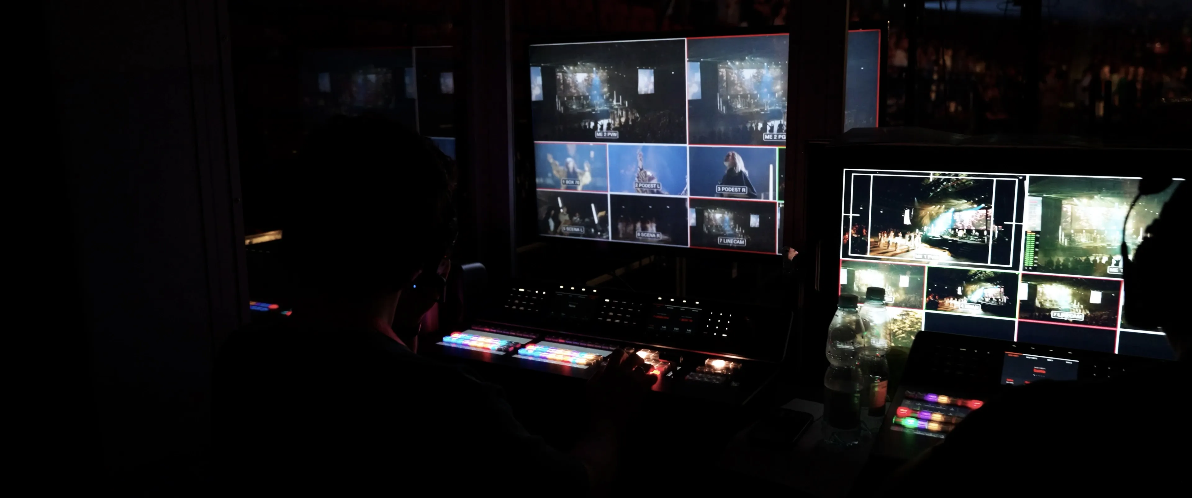 Person operating video production equipment with multiple screens displaying live event camera feeds in a dark control room.