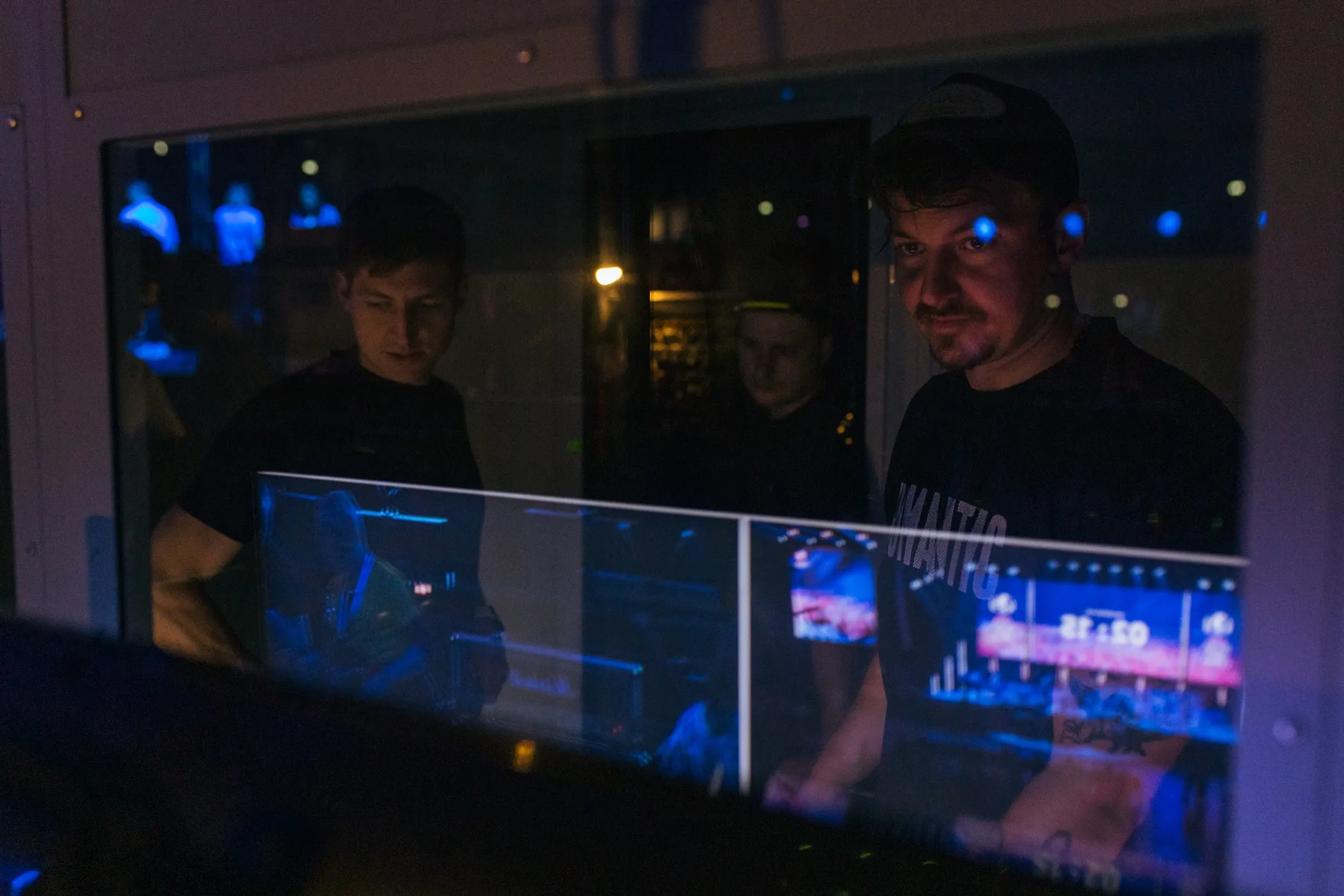 Three men focused on work behind a glass panel reflecting blue lights and screens in a dark room.