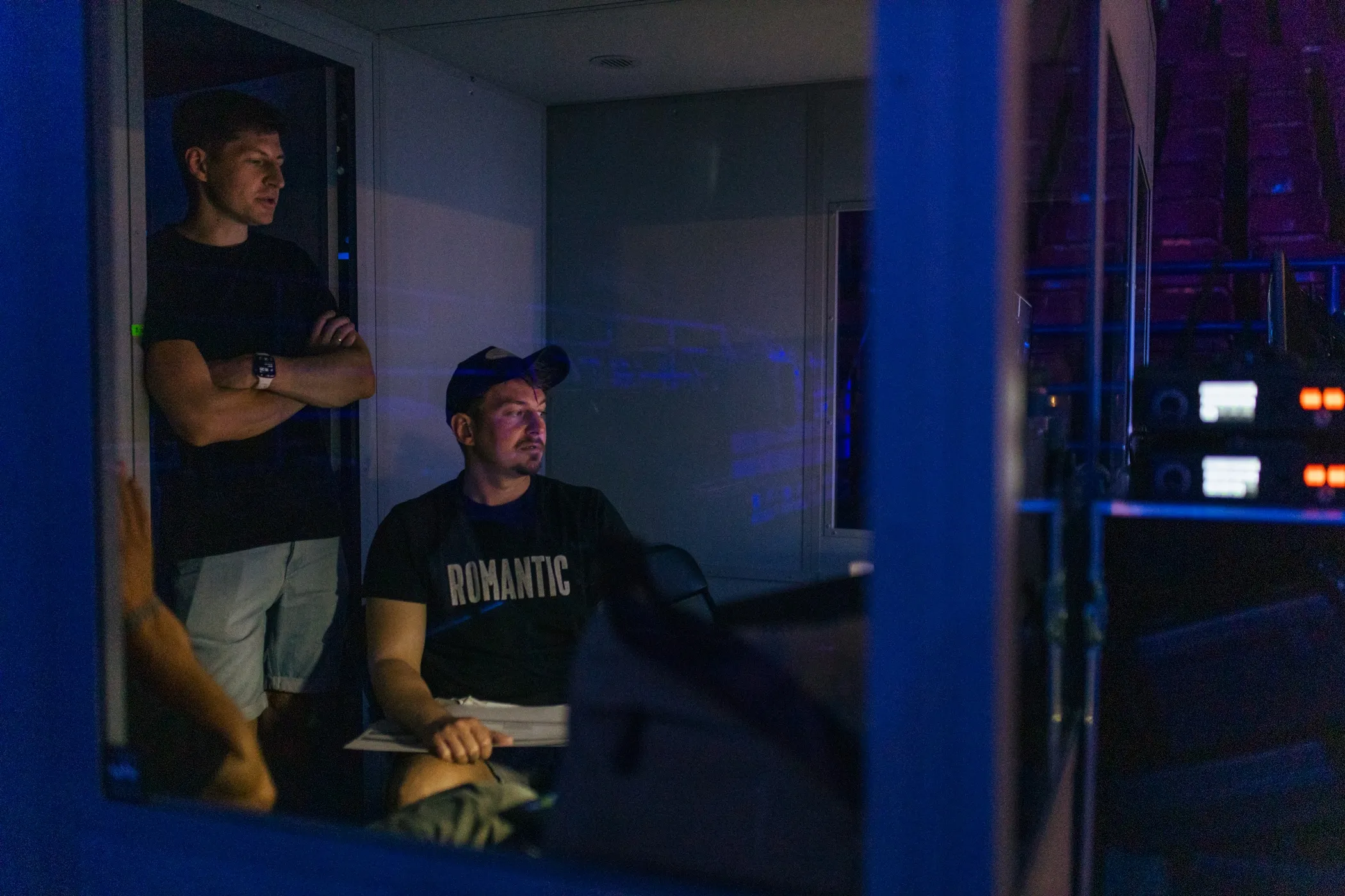 Two men in a dimly lit room, one standing with arms crossed and the other seated wearing a cap and a shirt with the word 'ROMANTIC'.