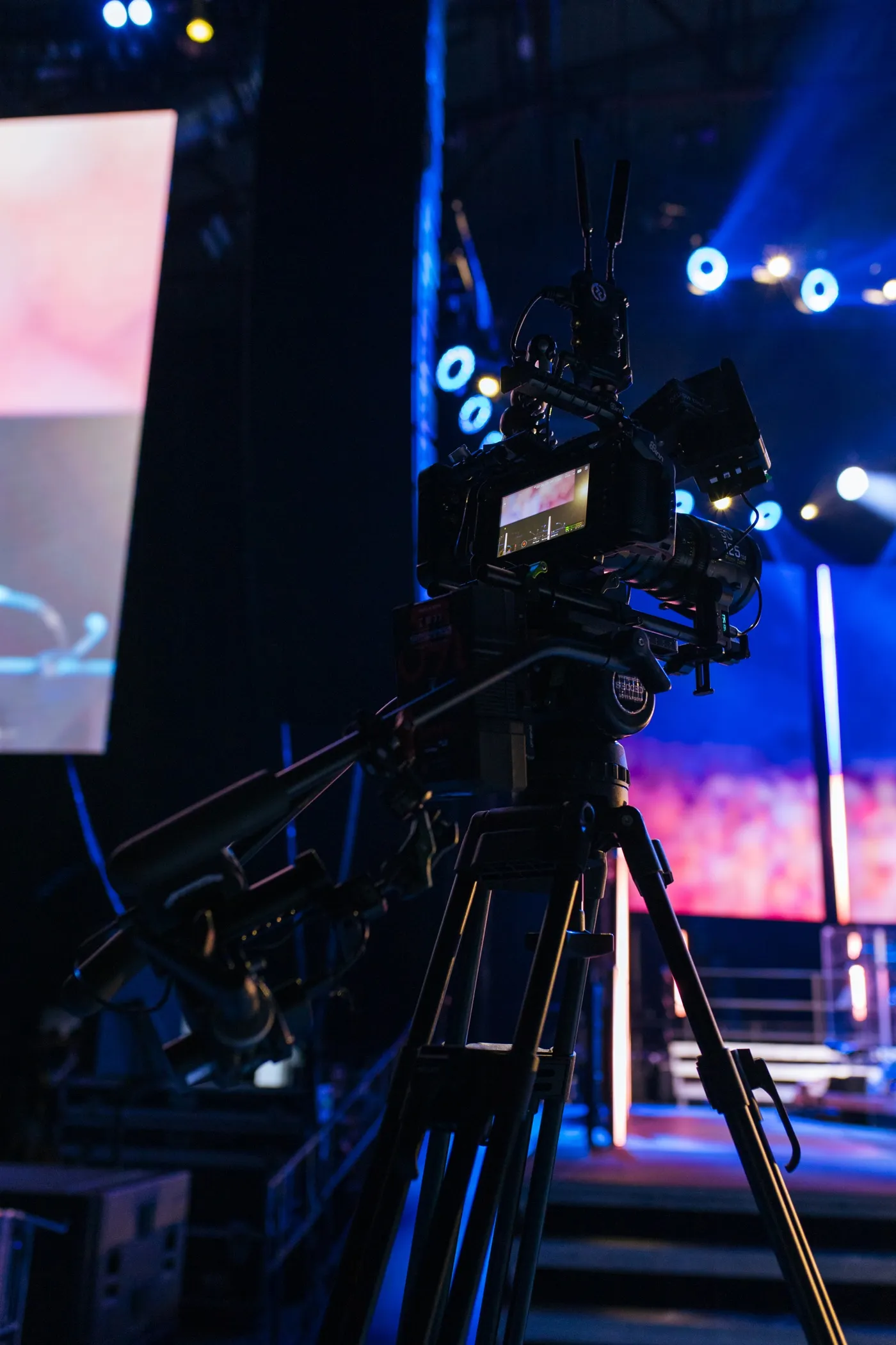 Profesjonalna kamera filmowa na statywie ustawiona w ciemnym studio z kolorowym oświetleniem na scenie.