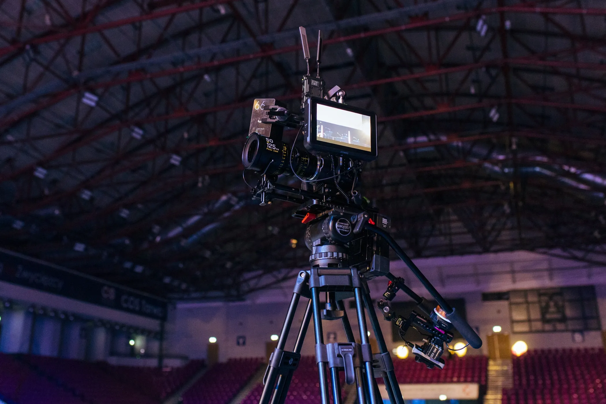 Profesjonalna kamera filmowa na statywie w hali z trybunami i oświetleniem.