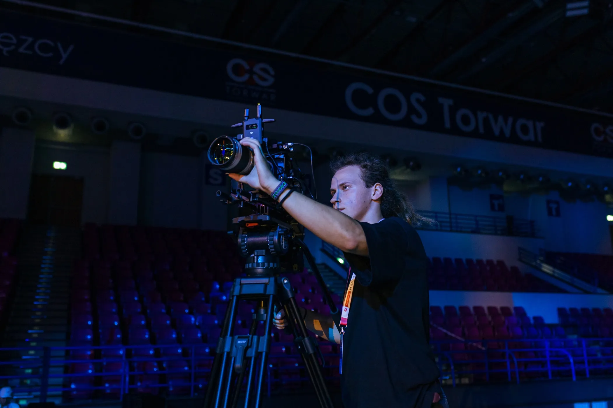 Młody operator kamery profesjonalnej ustawia obiektyw podczas nagrania na pustej trybunie hali COS Torwar.
