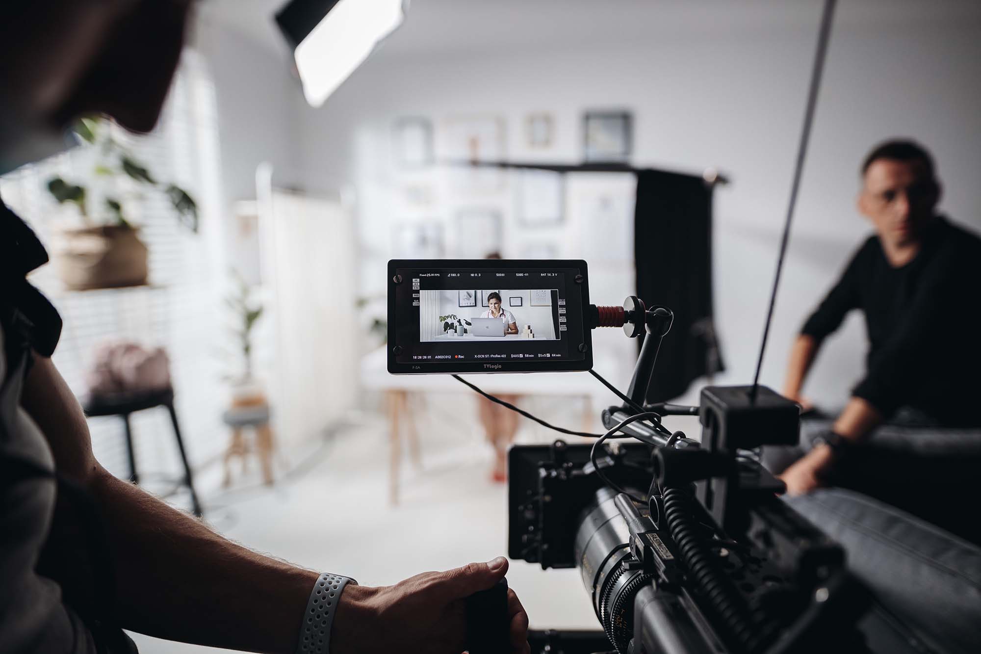 Camera operator from Ether Media filming a woman working on a laptop in a bright, modern office studio setup. Holding a Sony Venice camera.