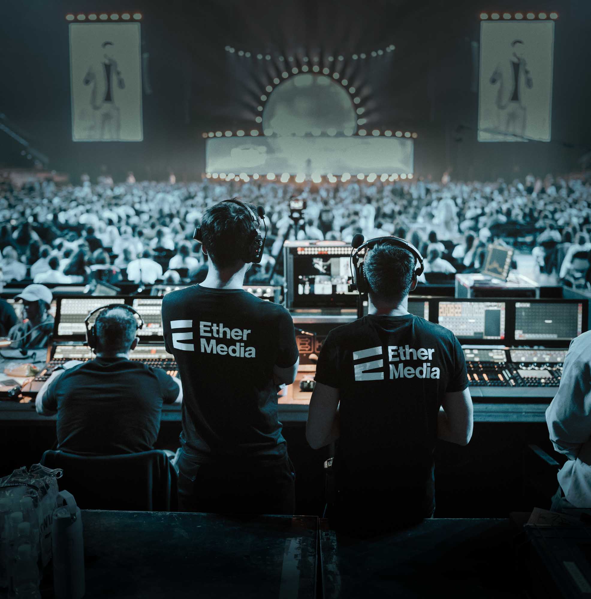 Crew wearing Ether Media shirts operate control panels at a live event with a large audience and stage in the background.