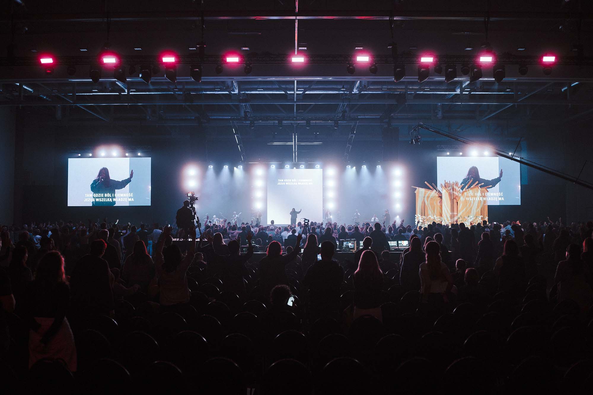A large crowd at an indoor concert or worship event with a band on stage, bright stage lights, and two large screens showing a singer and Polish lyrics.