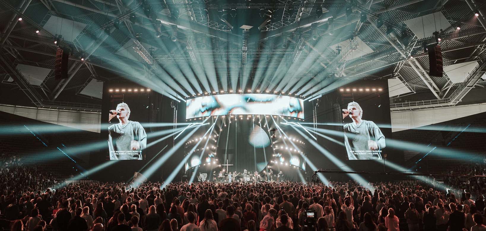 Large indoor concert with a crowd facing a brightly lit stage featuring a singer and musicians, with big screens displaying the singer.