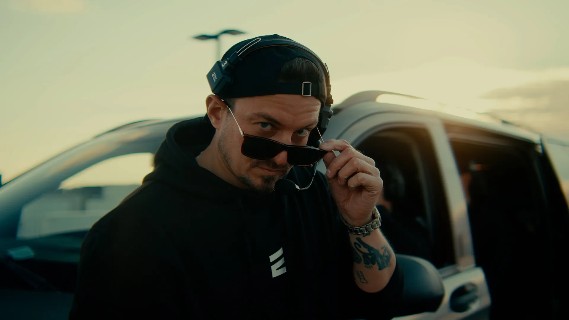 Mężczyzna w czarnej bluzie i czapce z mikrofonem na głowie zdejmuje okulary przeciwsłoneczne stojąc obok samochodu o zachodzie słońca.