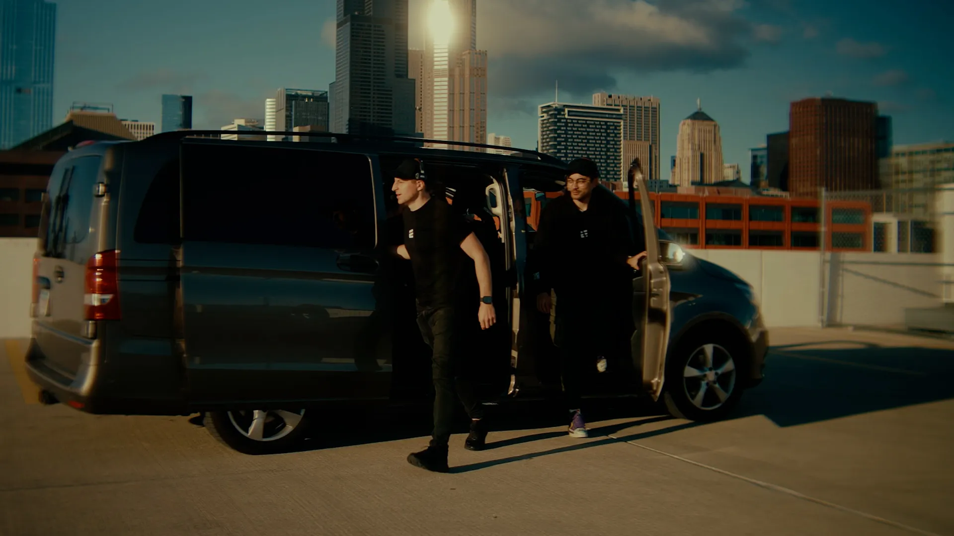 Dwóch mężczyzn w czarnych ubraniach wychodzi z ciemnego vana na tle miasta oświetlonego zachodzącym słońcem.
