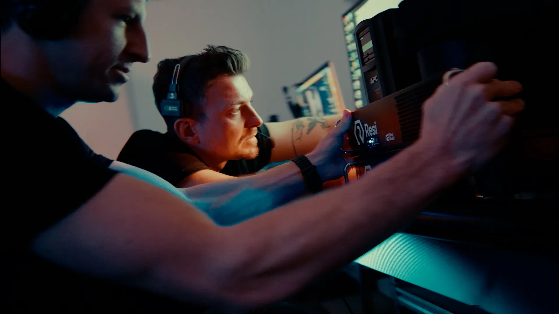 Dwaj mężczyźni pracujący z urządzeniem serwerowym w ciemnym pomieszczeniu z monitorami komputerowymi.