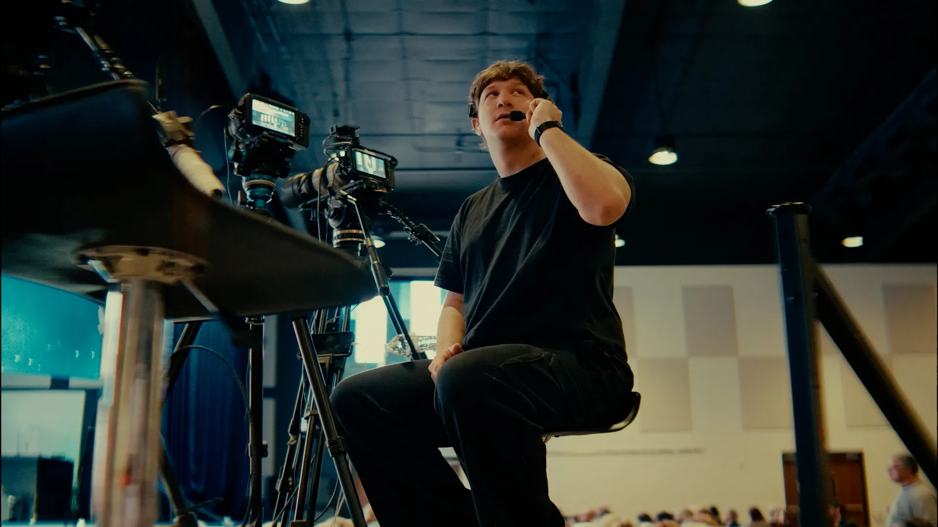 Mężczyzna w czarnej koszulce i słuchawkach z mikrofonem siedzi na stołku obok kamer w pomieszczeniu z widownią.