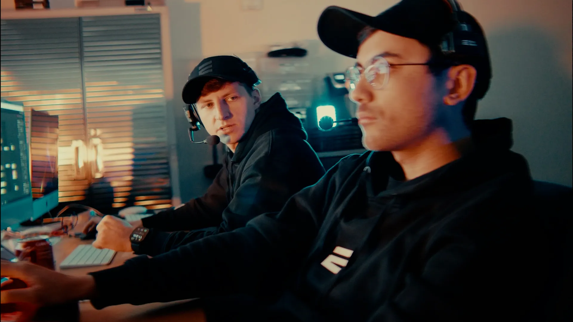 Two young men wearing headsets and black hoodies working together at computer desks with multiple monitors in a dimly lit room.