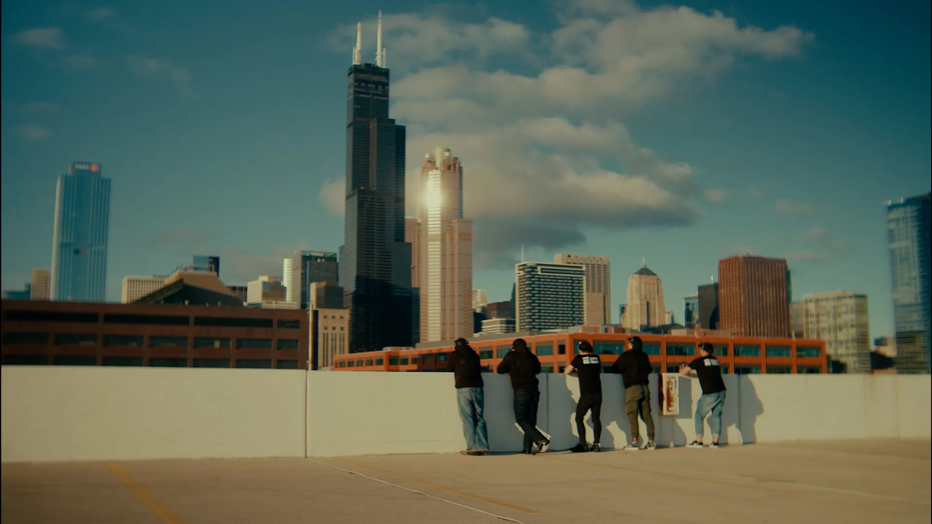 Grupa pięciu osób opiera się o barierę na dachu parkingu z widokiem na panoramę miasta z wysokimi wieżowcami, w tym charakterystycznym ciemnym budynkiem Willis Tower.