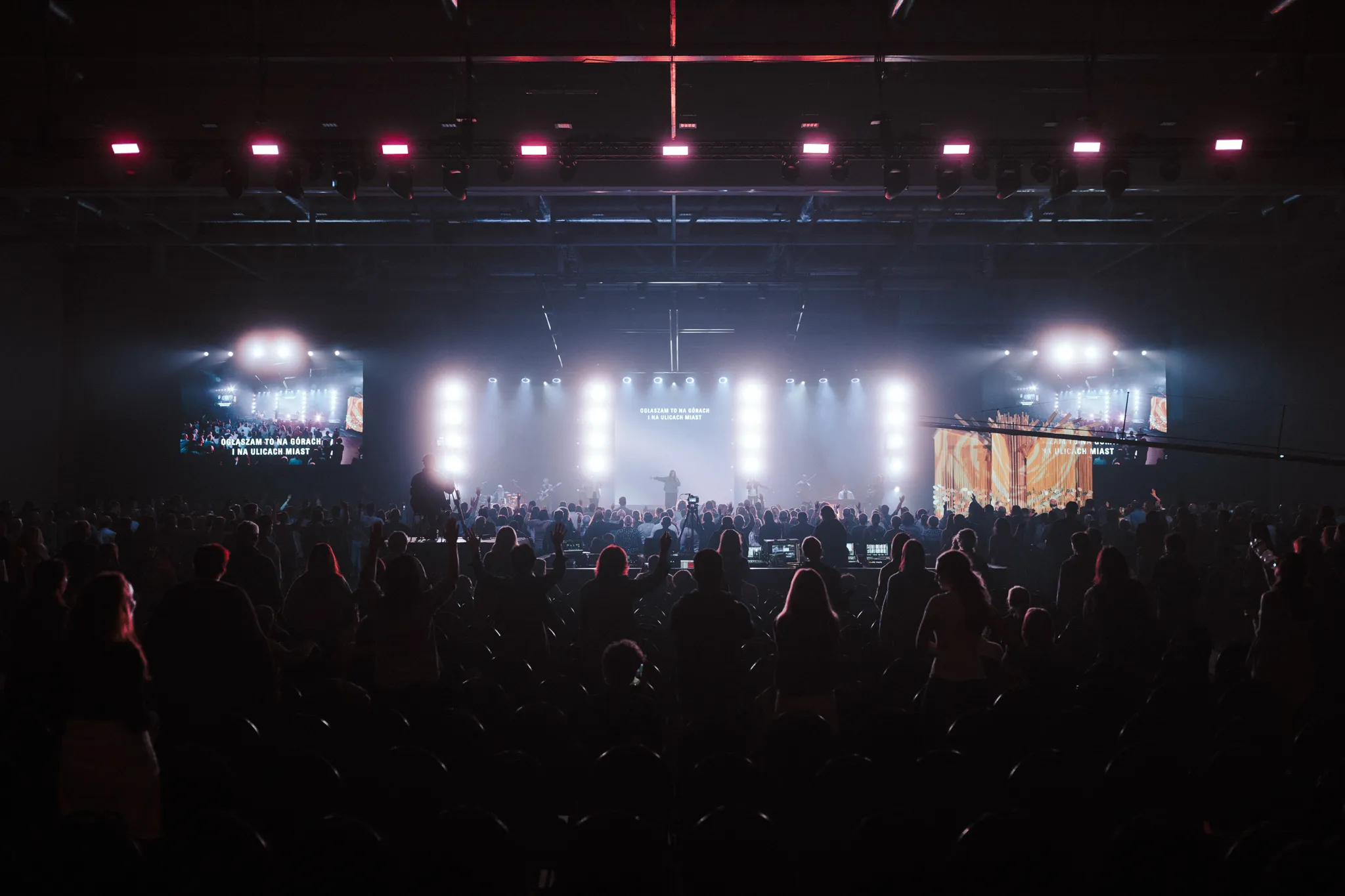 Audience standing and watching a brightly lit stage with performers and large LED screens inside a concert venue.