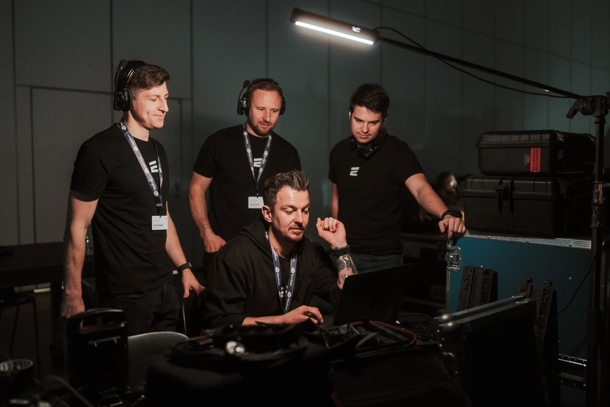 Four men with headsets gathered around a seated man working on a laptop in a dimly lit control room.