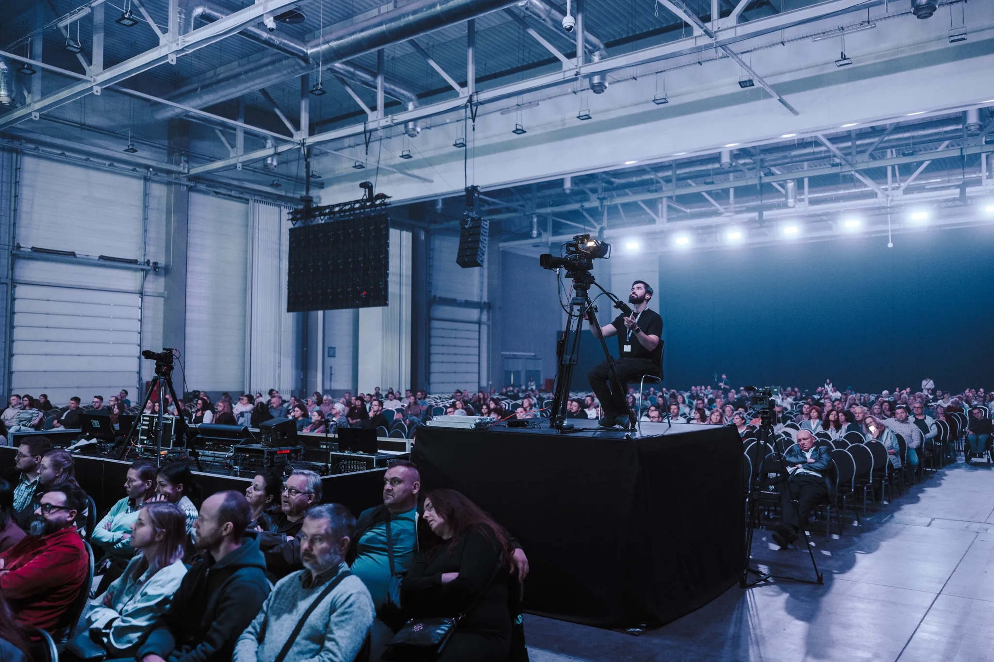 Operator kamerzysta siedzi na podwyższeniu i nagrywa dużą publiczność siedzącą w hali konferencyjnej.
